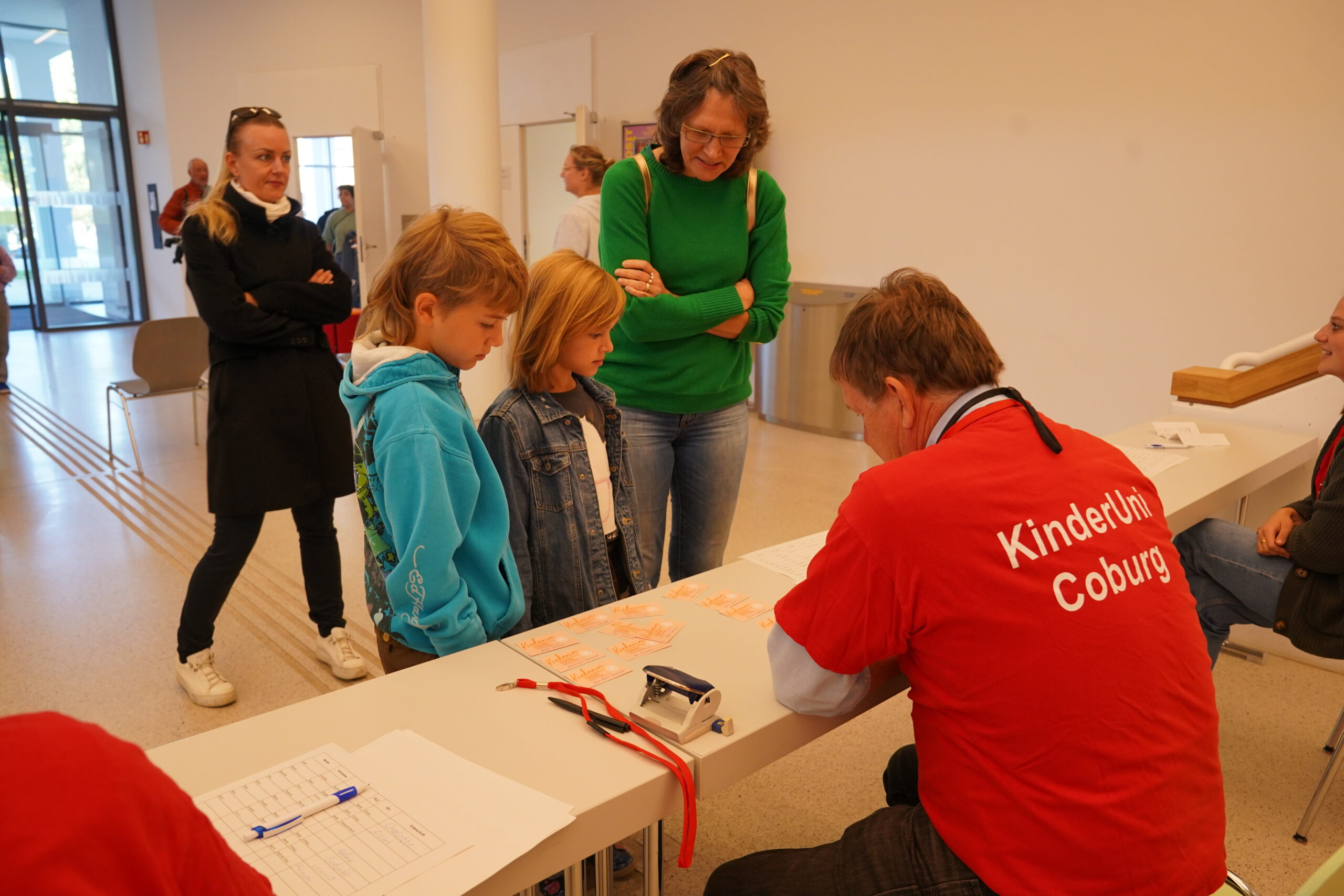 Zwei Kinder der KinderUni stehen an einem Tisch und schauen sich Anmeldekarten an, während ein Mann in einem roten Hemd die Daten dieser Stammgäste erfasst. at Hochschule Coburg