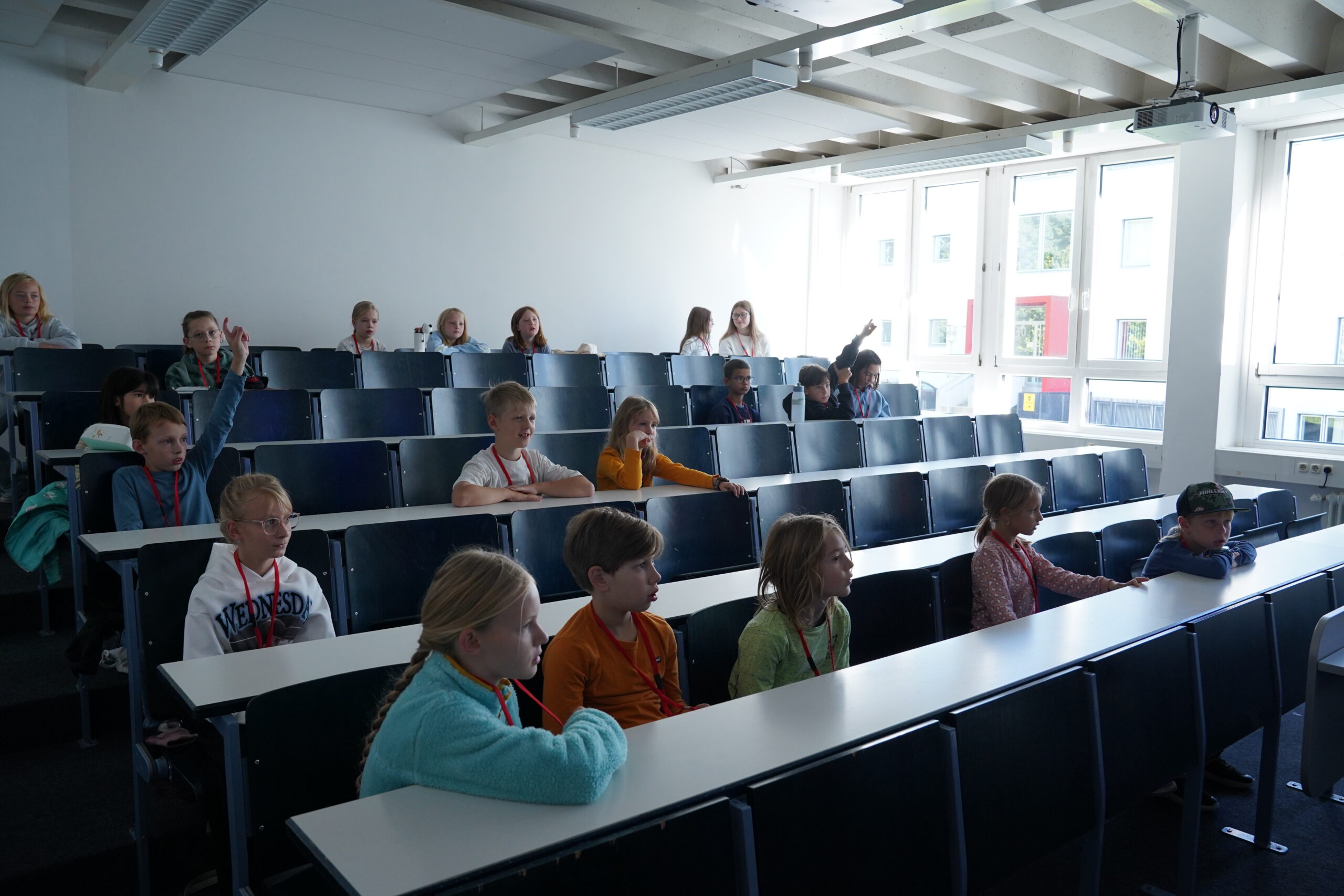 Die Kinder sitzen an Schreibtischen in einem stufenförmig angeordneten Hörsaal der KinderUni, wobei einige die Hand heben. at Hochschule Coburg