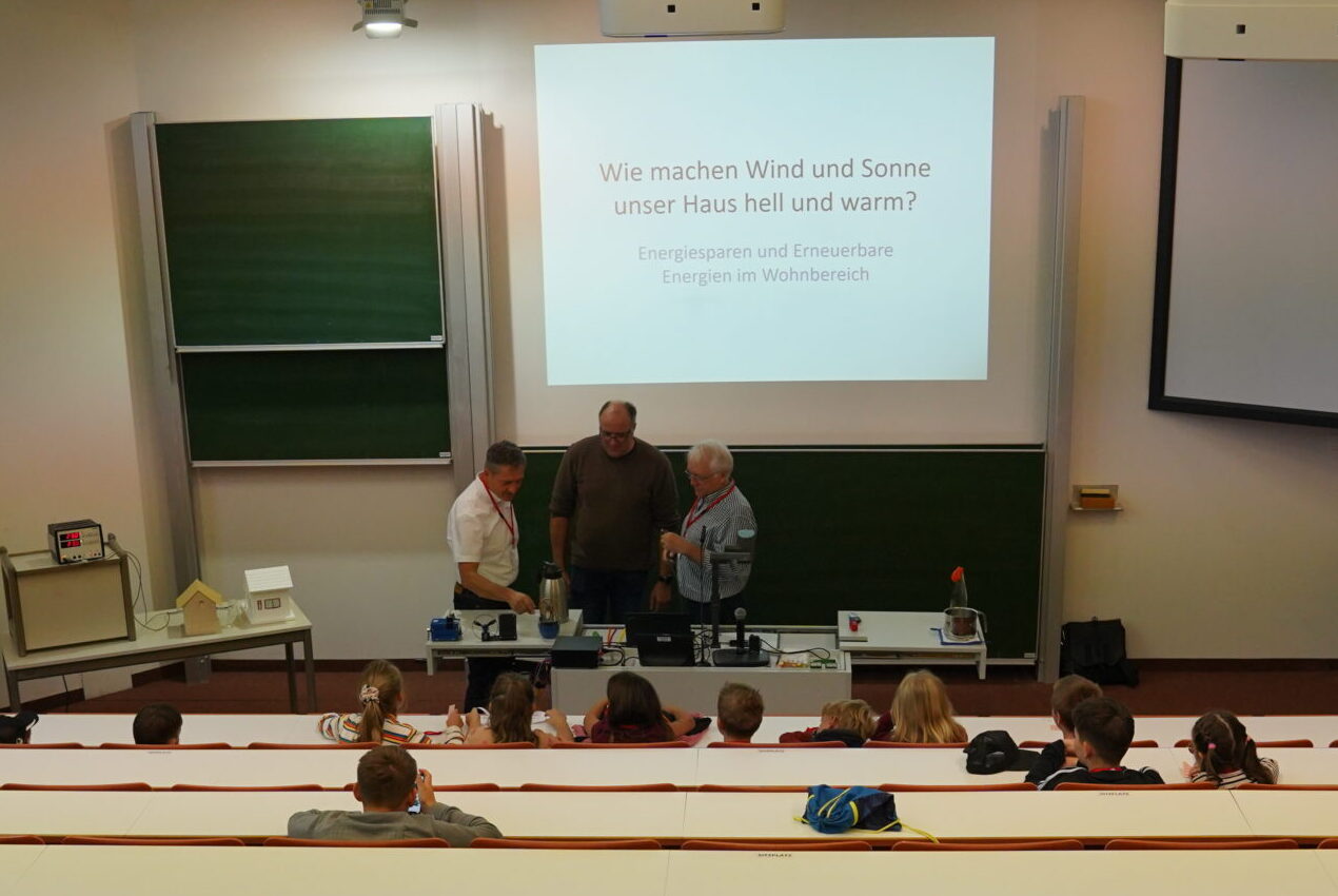 Drei Erwachsene führen in einem Hörsaal für Kinder eine naturwissenschaftliche Vorführung im Rahmen der „KinderUni“ durch, wobei auf einer Leinwand eine deutsche Präsentation zum Thema erneuerbare Energien mit dem Titel „Einmal ist keinmal“ gezeigt wird. at Hochschule Coburg