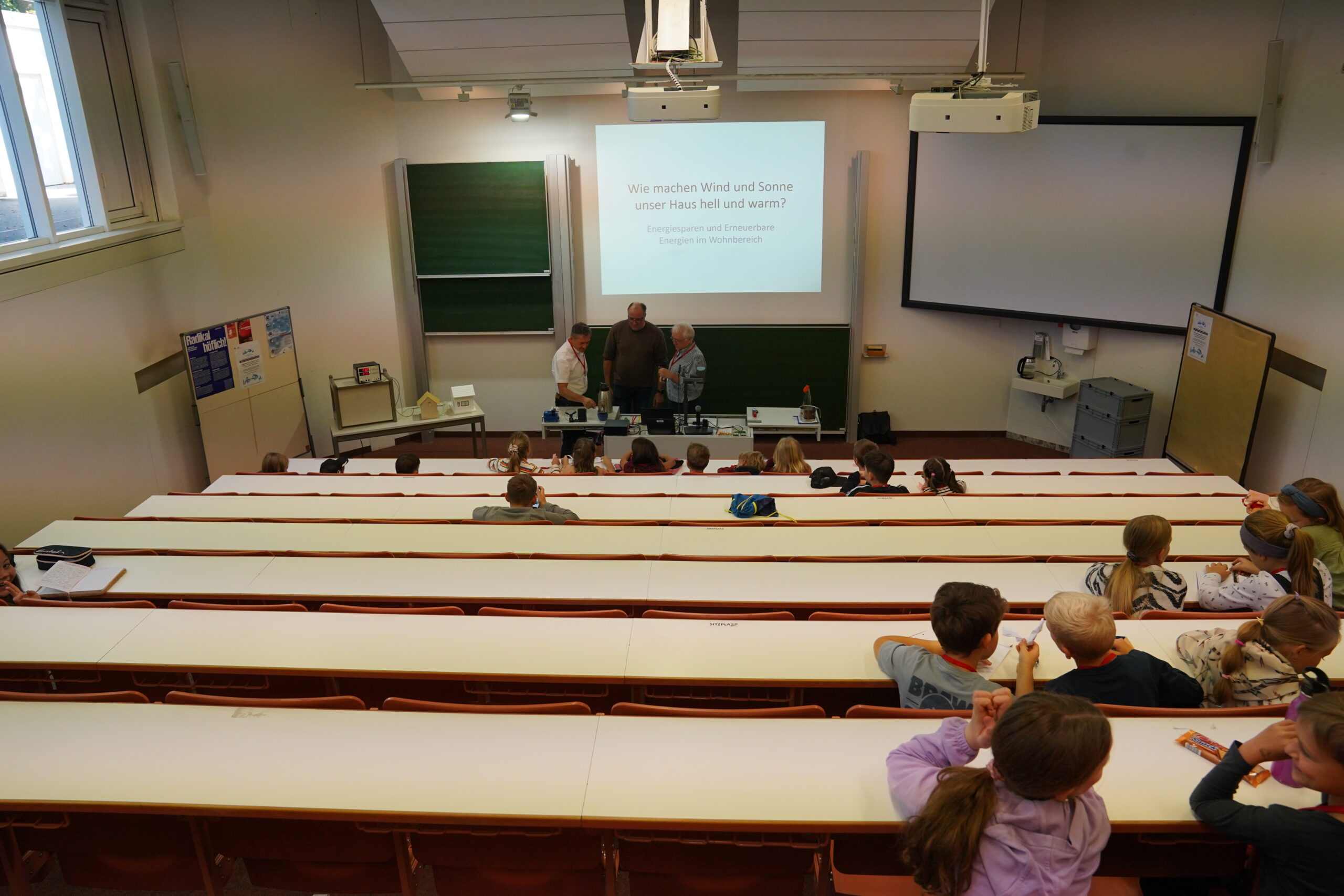 Drei Erwachsene führen in einem Hörsaal für Kinder eine naturwissenschaftliche Vorführung im Rahmen der „KinderUni“ durch, wobei auf einer Leinwand eine deutsche Präsentation zum Thema erneuerbare Energien mit dem Titel „Einmal ist keinmal“ gezeigt wird. at Hochschule Coburg
