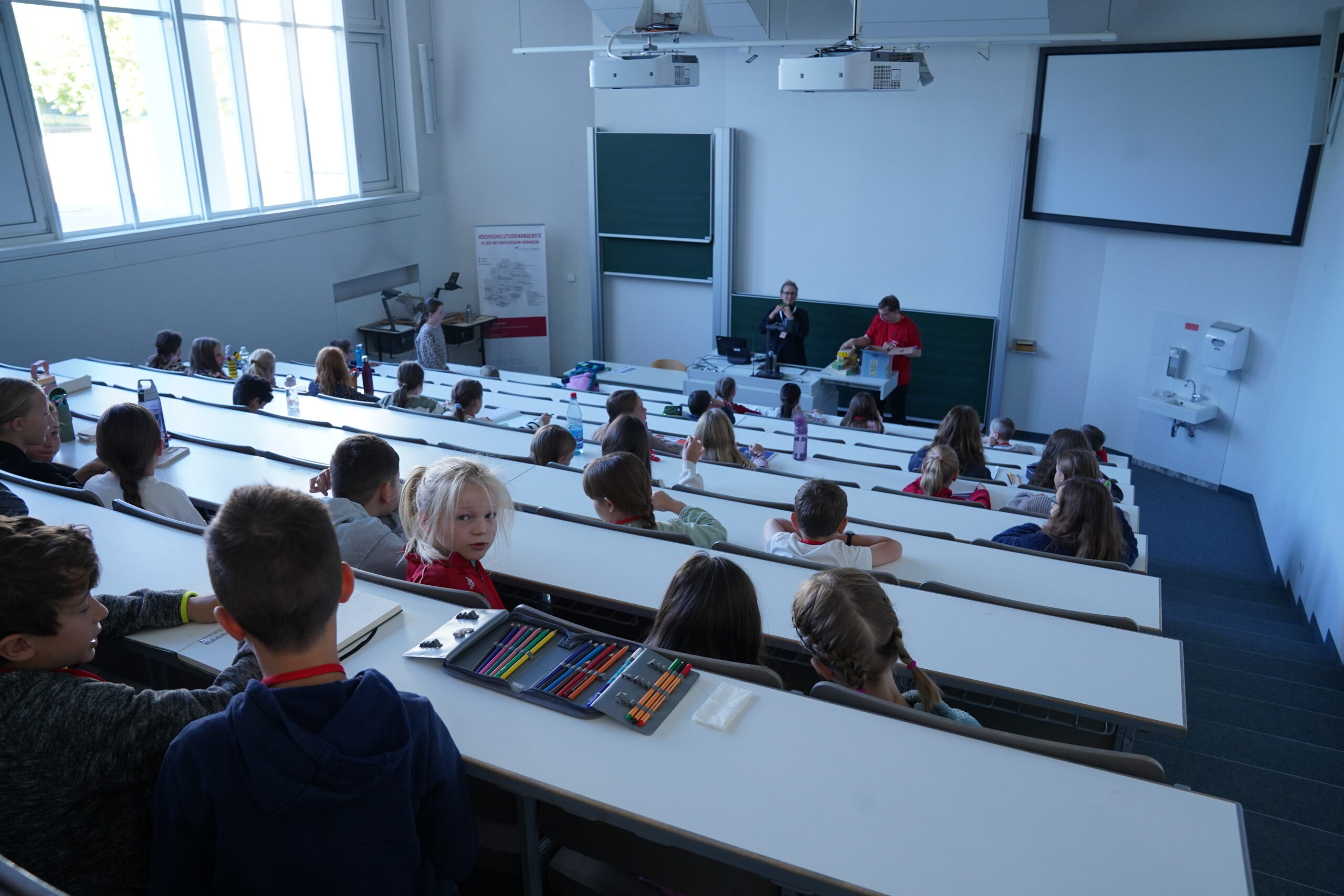 Die Studierenden sitzen in einem Hörsaal der KinderUni, während zwei Referenten vorne an einer Tafel stehen. at Hochschule Coburg