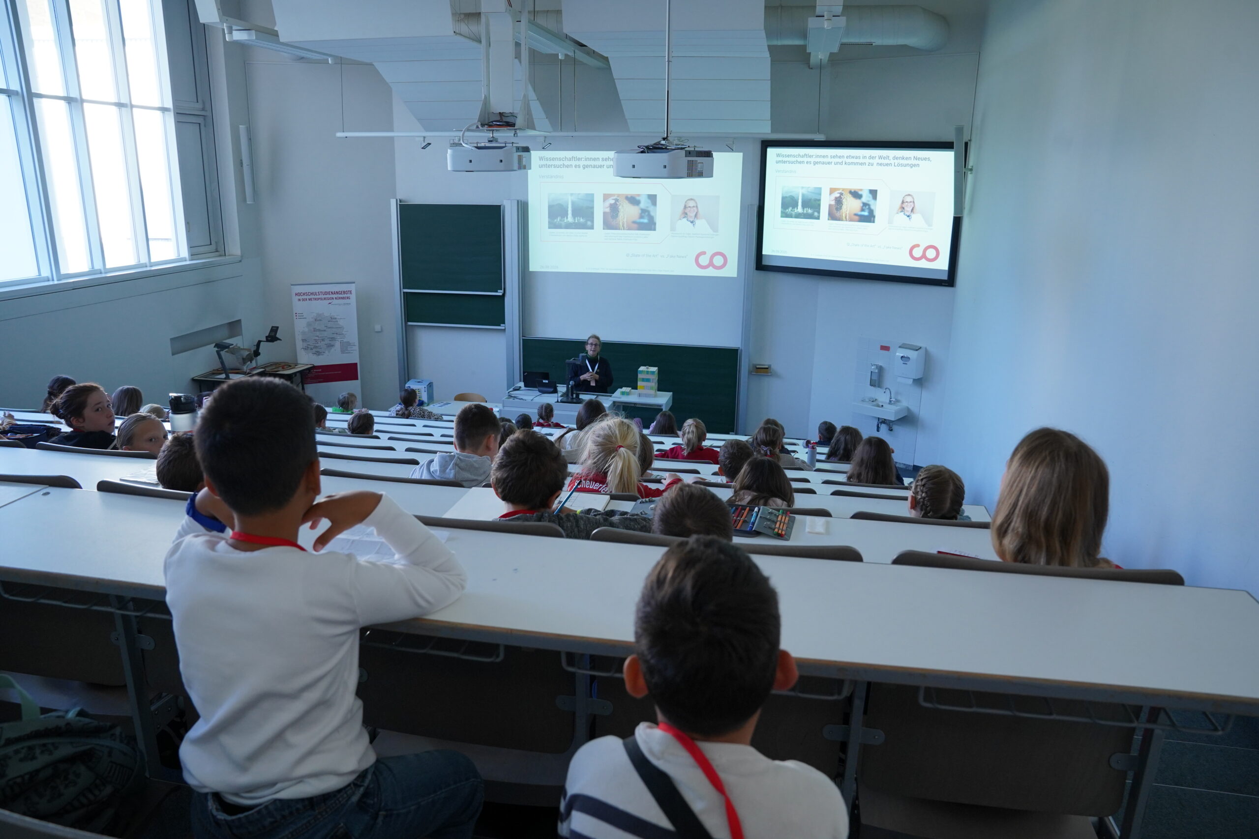 Ein Dozent hält vor den Teilnehmern der KinderUni in einem stufenförmig angelegten Hörsaal mit zwei Projektionsleinwänden einen Vortrag über wissenschaftliche Forschung – eine Veranstaltung, bei der ein Besuch nie ausreicht. at Hochschule Coburg
