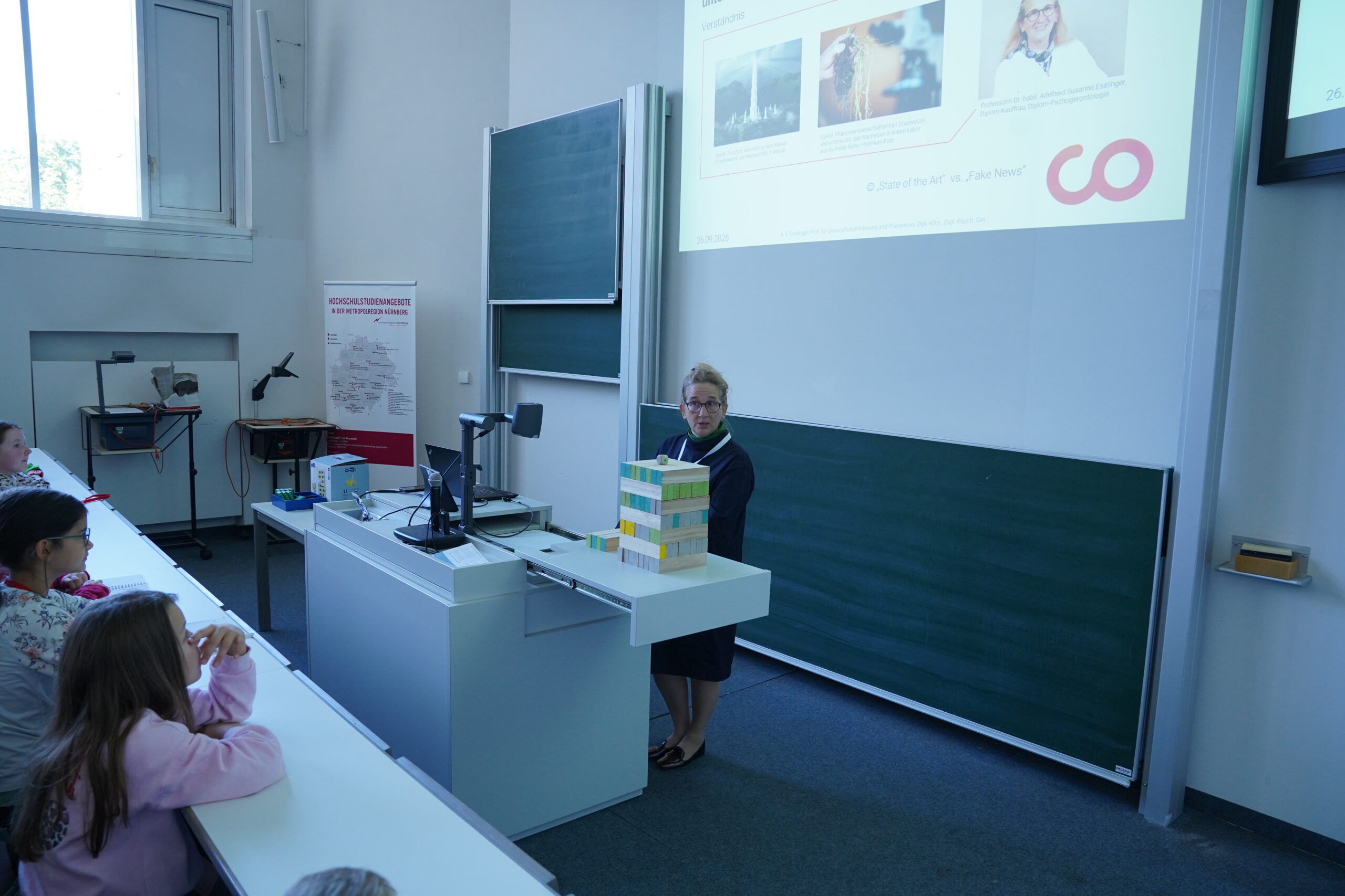 Ein Dozent steht vorne im Unterrichtsraum der KinderUni neben einer Projektionswand, während die Stammgäste an ihren Tischen sitzen. at Hochschule Coburg