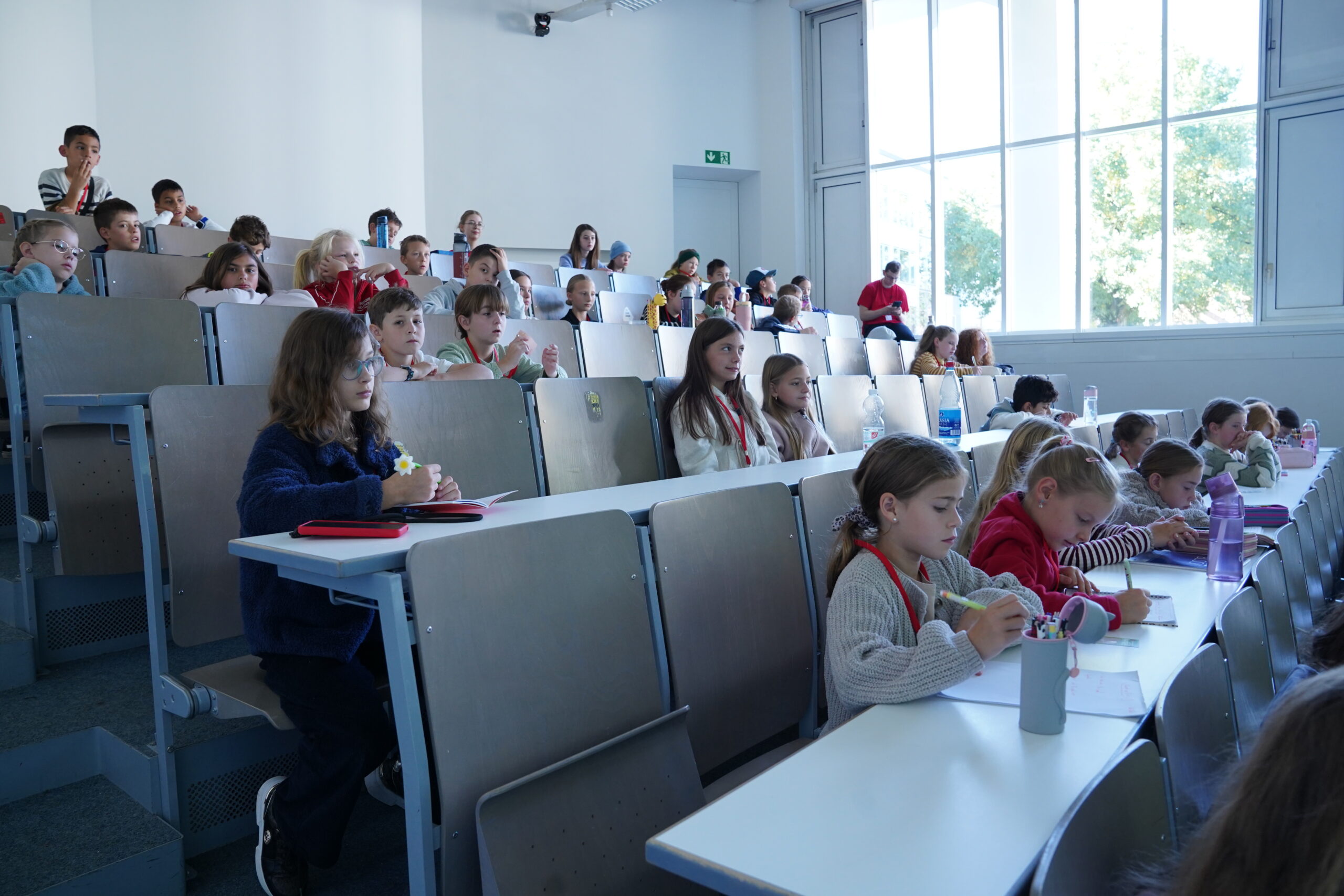 Die Studierenden nehmen an einer Vorlesung der KinderUni teil und sitzen an Tischen in einem stufenförmig angelegten Hörsaal, in dem viele von ihnen Stammgäste sind. at Hochschule Coburg