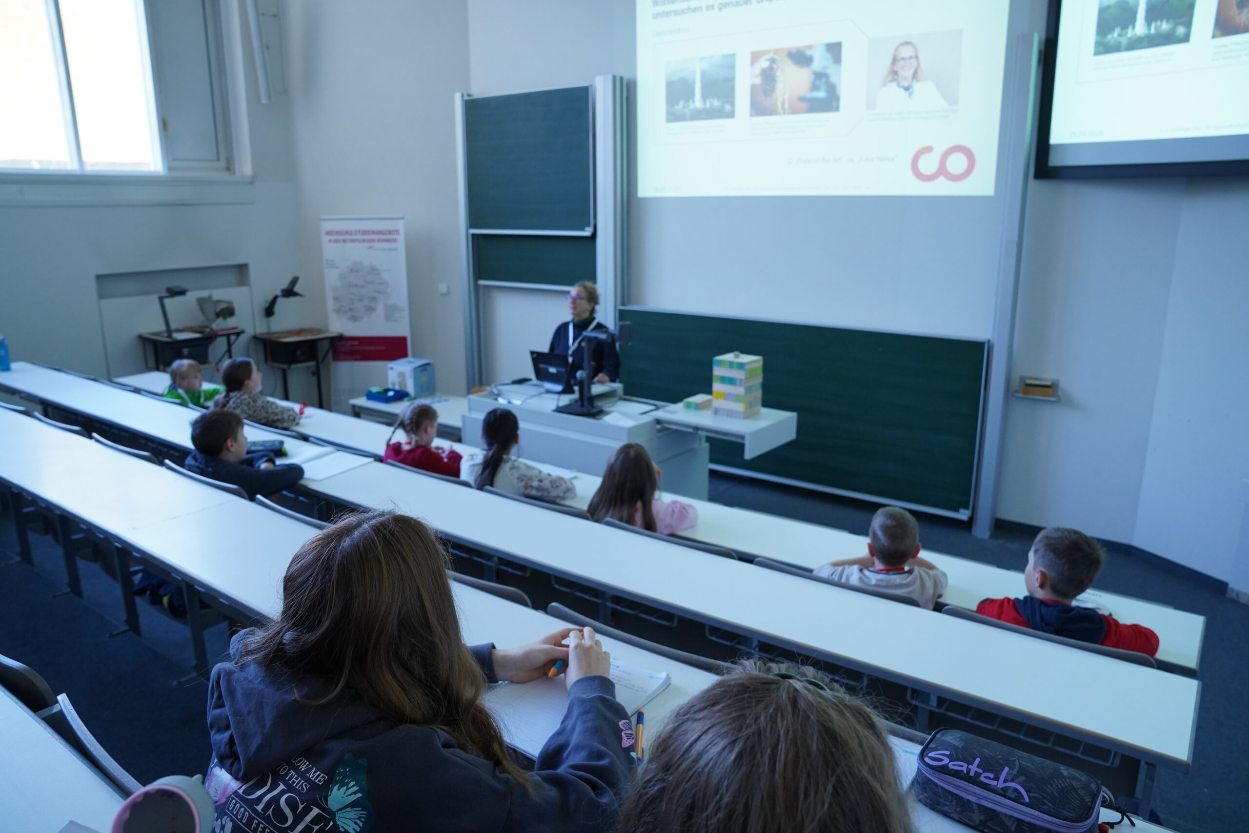 Ein Lehrer hält vor einer Gruppe von „KinderUni“-Studenten einen Vortrag, die an Schreibtischen in einem Hörsaal sitzen, wo sich zahlreiche Stammgäste versammelt haben, um auf die Leinwand zu schauen. at Hochschule Coburg
