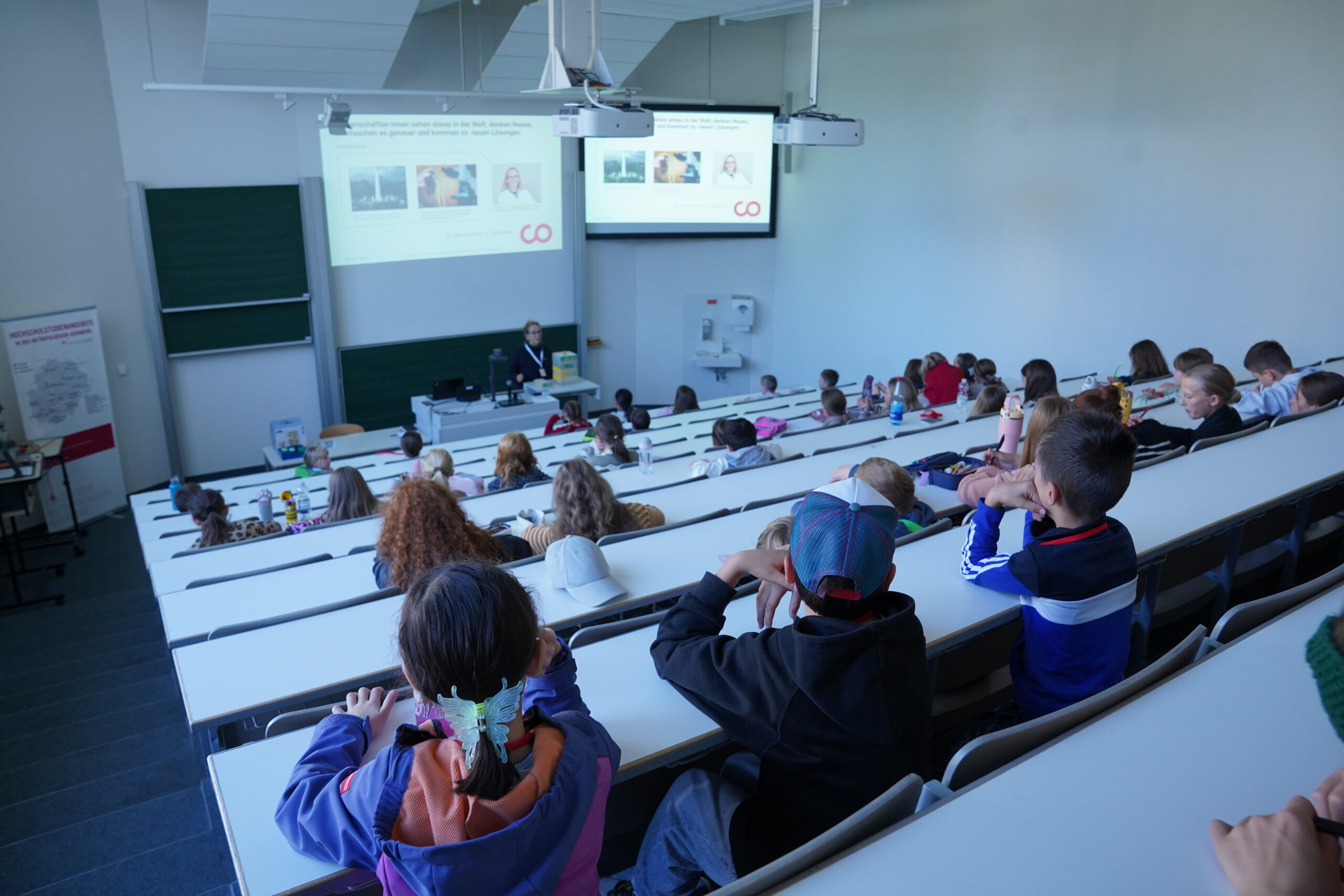 Stammgäste und andere Studierende sitzen in einem stufenförmig angeordneten Hörsaal der Universität und blicken auf einen Redner sowie auf Präsentationsbildschirme. at Hochschule Coburg