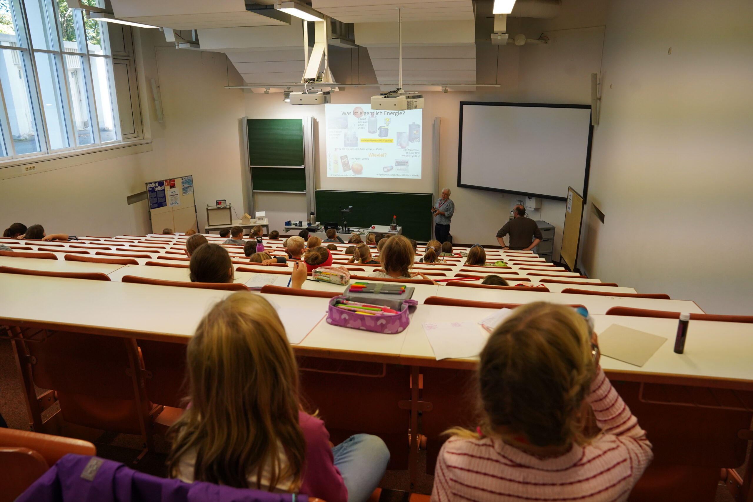 Ein Dozent steht vorne in einem stufenförmig angelegten Hörsaal der KinderUni, wo eine Mischung aus Neulingen und Stammgästen an Schultischen sitzt, die auf eine Leinwand ausgerichtet sind. at Hochschule Coburg