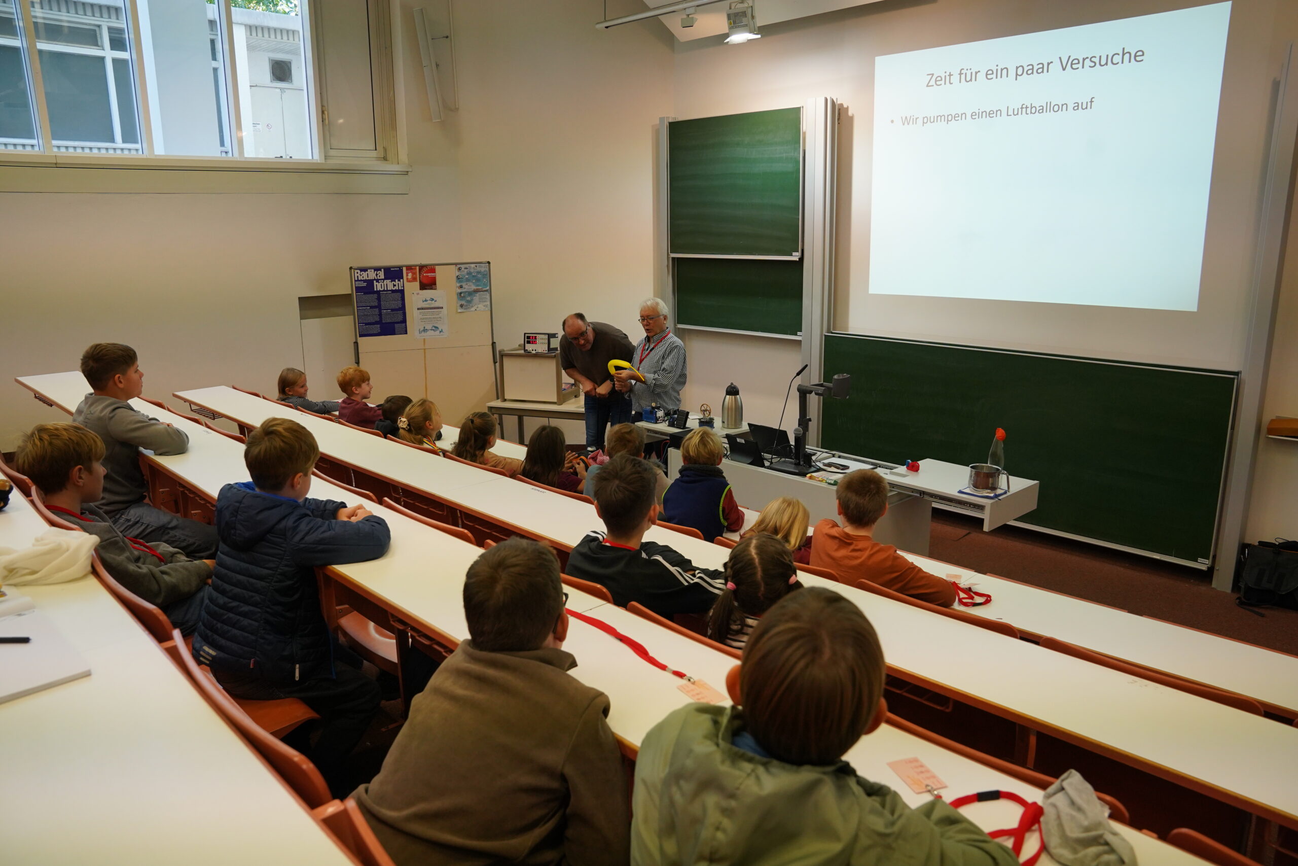 Zwei KinderUni-Dozenten führen in einem Hörsaal ein Ballon-Experiment durch, während auf einer Leinwand ein deutscher Text eingeblendet wird – was einmal mehr beweist: „Einmal ist keinmal.“ at Hochschule Coburg