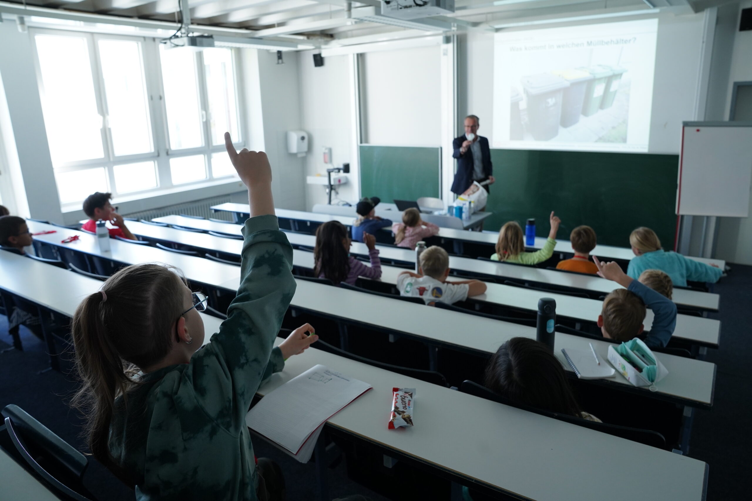 Ein Dozent steht vor einer KinderUni-Gruppe, während die Schüler auf den gestuften Sitzplätzen die Hand heben, um eine auf die Leinwand projizierte Frage zu beantworten. at Hochschule Coburg