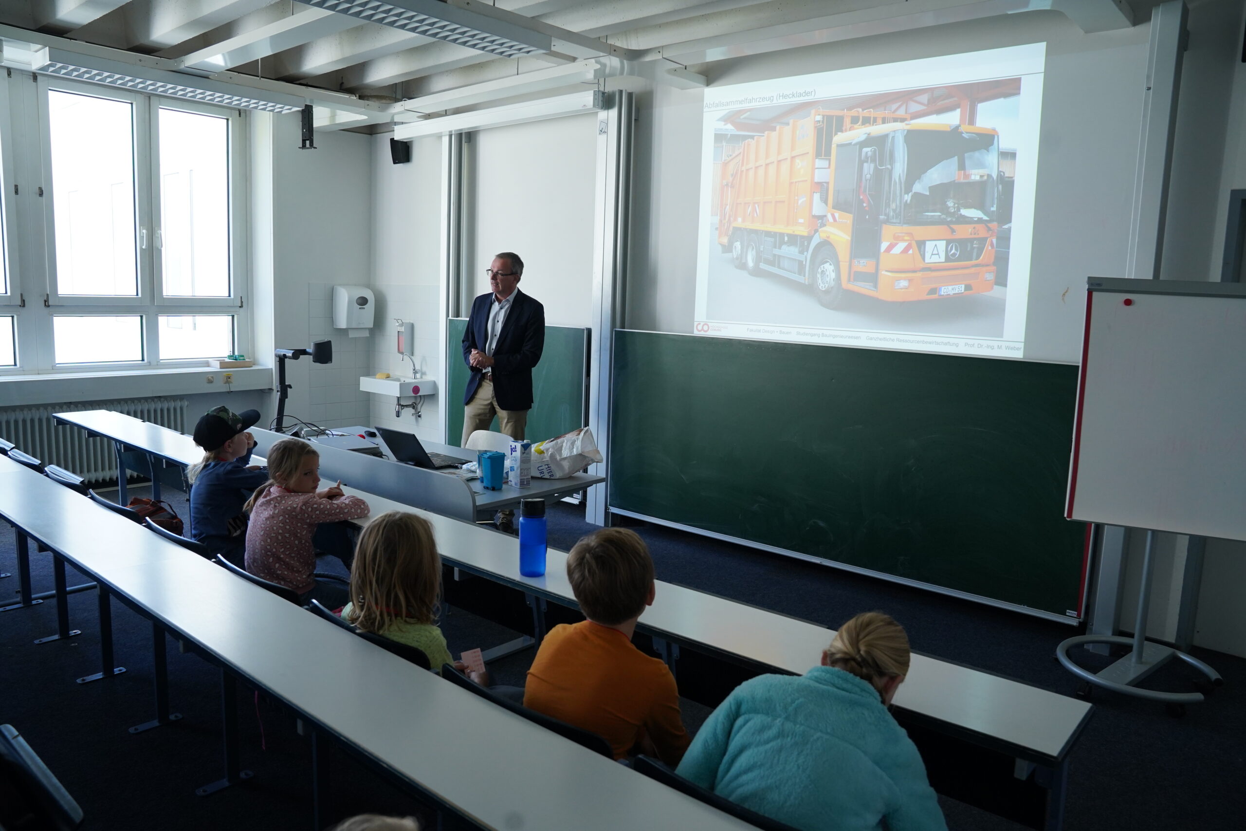 Ein Mann im Anzug hält vor einer Gruppe von Stammgästen an der KinderUni in einem Klassenzimmer einen Vortrag, während an der Wand das Bild eines orangefarbenen Müllwagens projiziert wird. at Hochschule Coburg