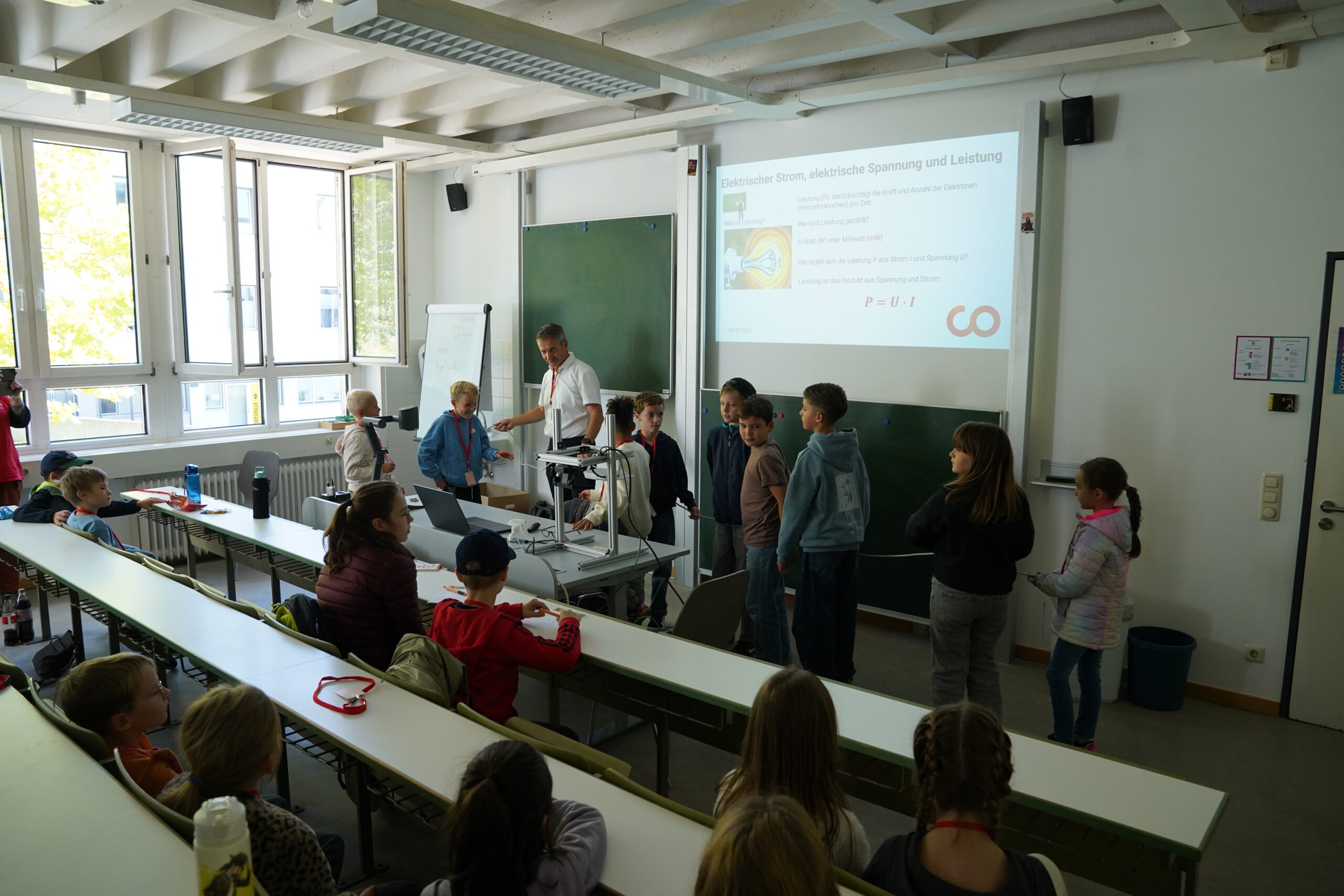 Ein Lehrer führt für die Teilnehmer der KinderUni in einem Klassenzimmer ein Experiment vor, während im Hintergrund auf einer Leinwand physikalische Notizen zum Thema elektrische Leistung zu sehen sind und Stammgäste den Unterricht beobachten. at Hochschule Coburg