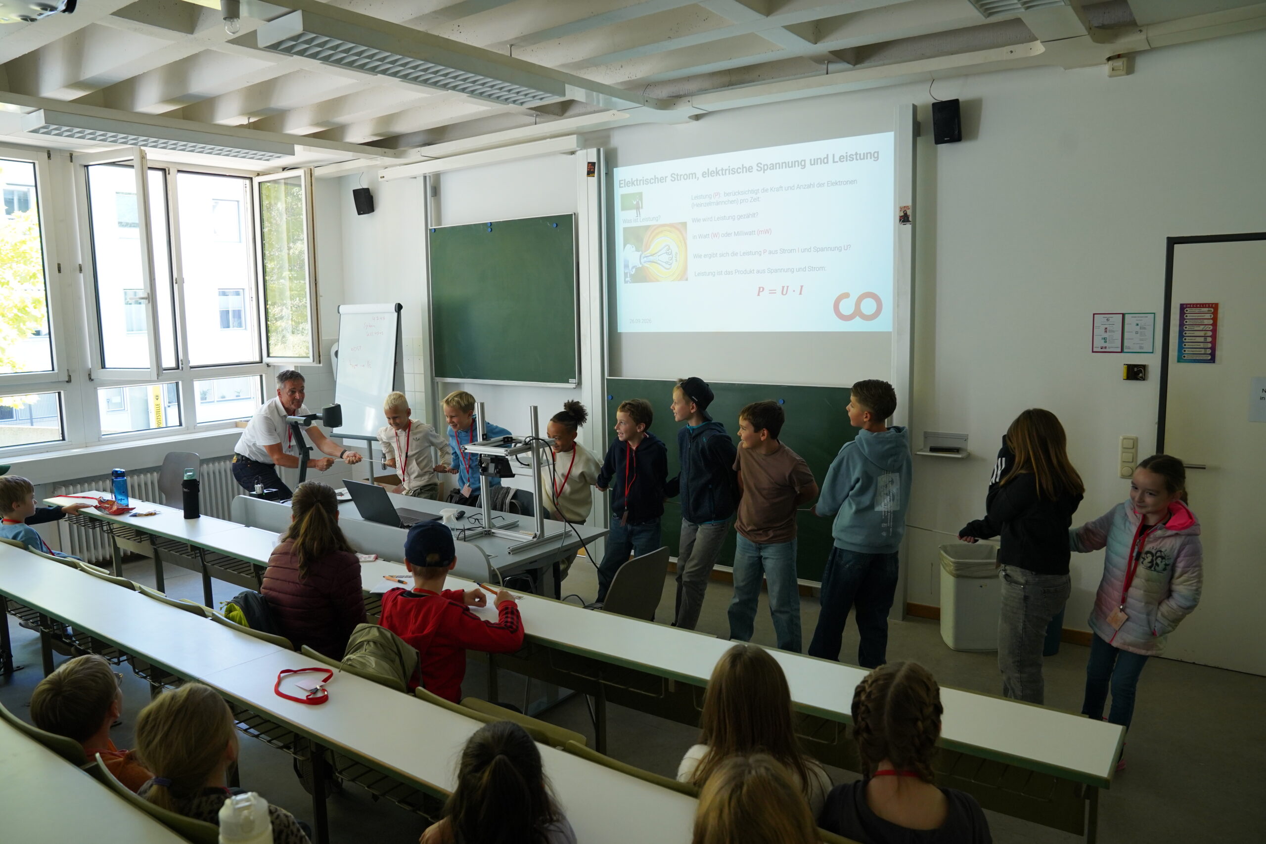 Ein Lehrer führt einer Gruppe von „KinderUni“-Teilnehmern ein elektrisches Experiment vor, während auf einem Projektor eine Unterrichtseinheit zum Thema Spannung und Leistung gezeigt wird. at Hochschule Coburg