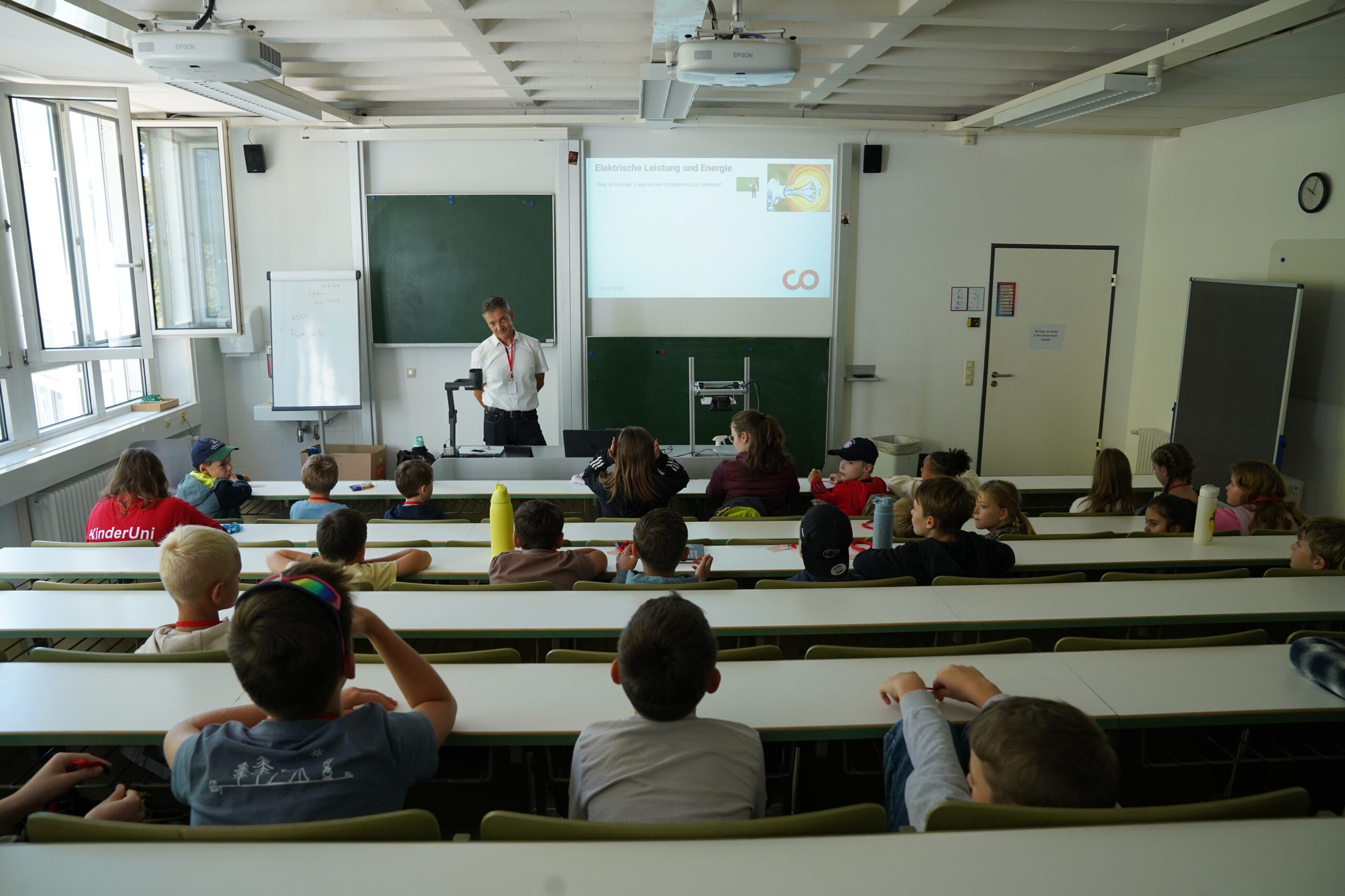 Ein Lehrer hält vor einem Raum voller aufmerksamer Stammgäste der KinderUni einen Vortrag über elektrische Leistung und Energie. at Hochschule Coburg