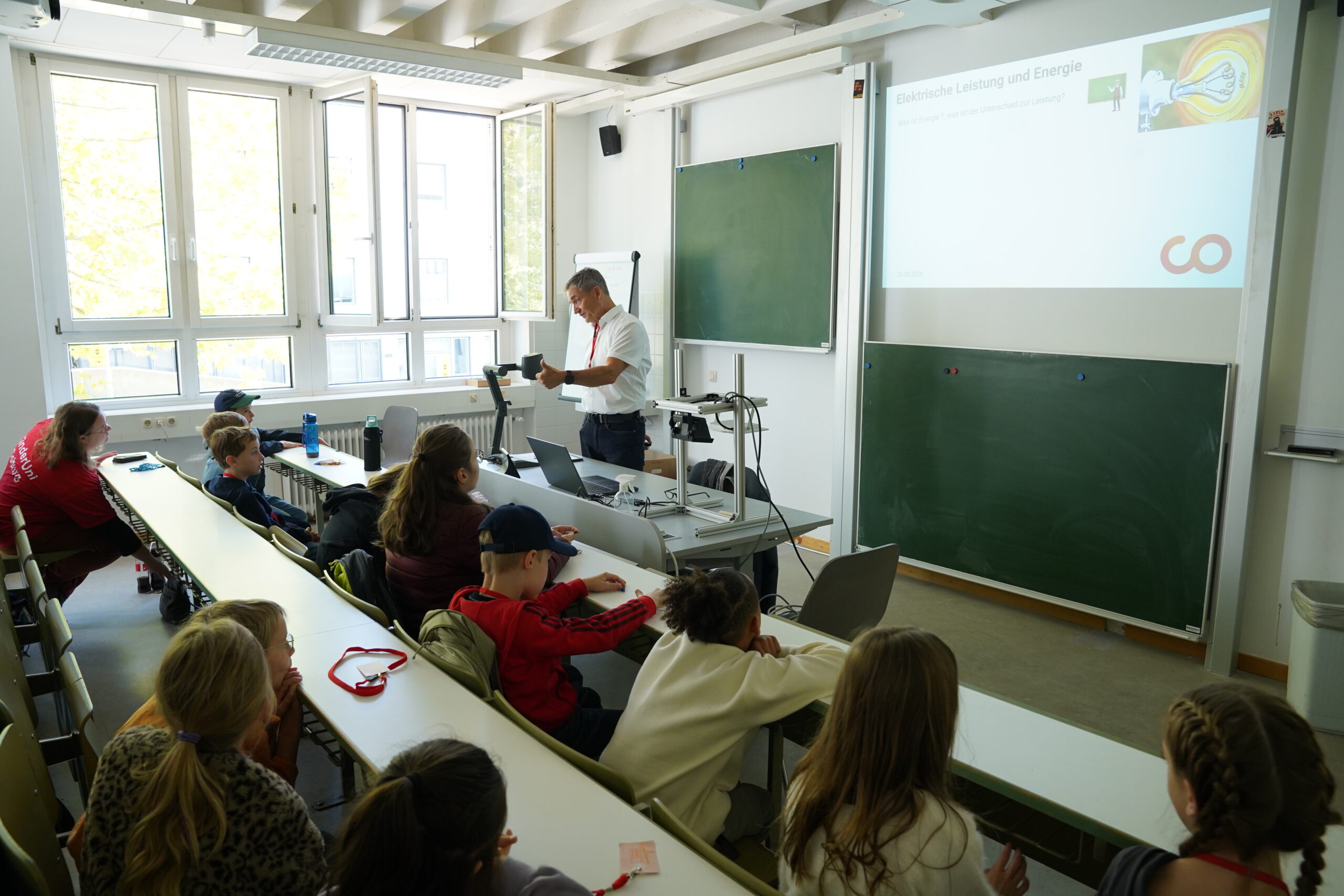 Ein männlicher Dozent zeigt den Teilnehmern der KinderUni an ihren Tischen, die auf eine Projektionswand mit der Aufschrift „Elektrische Leistung und Energie“ gerichtet sind, ein Gerät. at Hochschule Coburg
