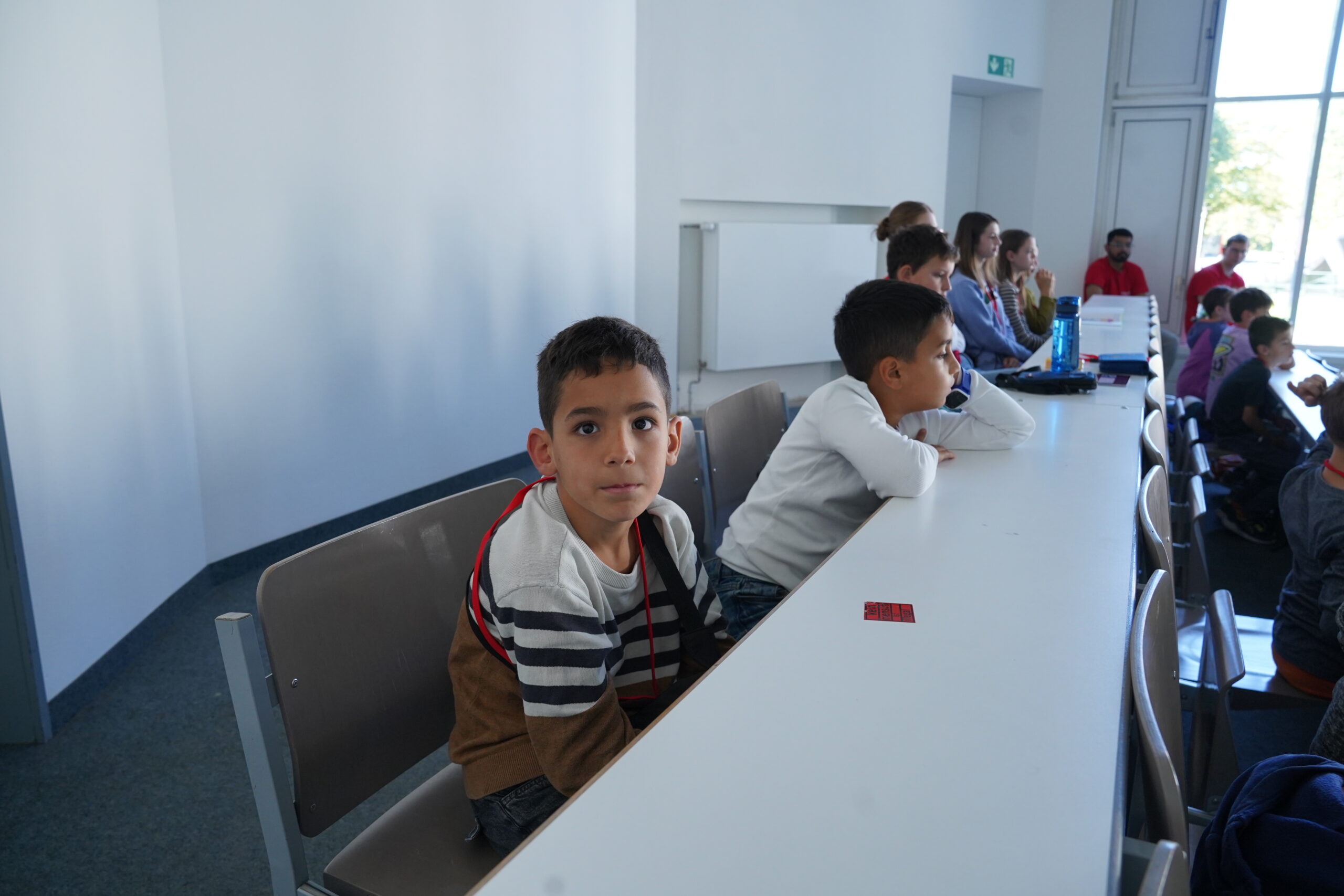 Ein Junge sitzt an einem Schreibtisch in der KinderUni und blickt in die Kamera, während andere Stammgäste hinter ihm in einer Reihe sitzen. at Hochschule Coburg