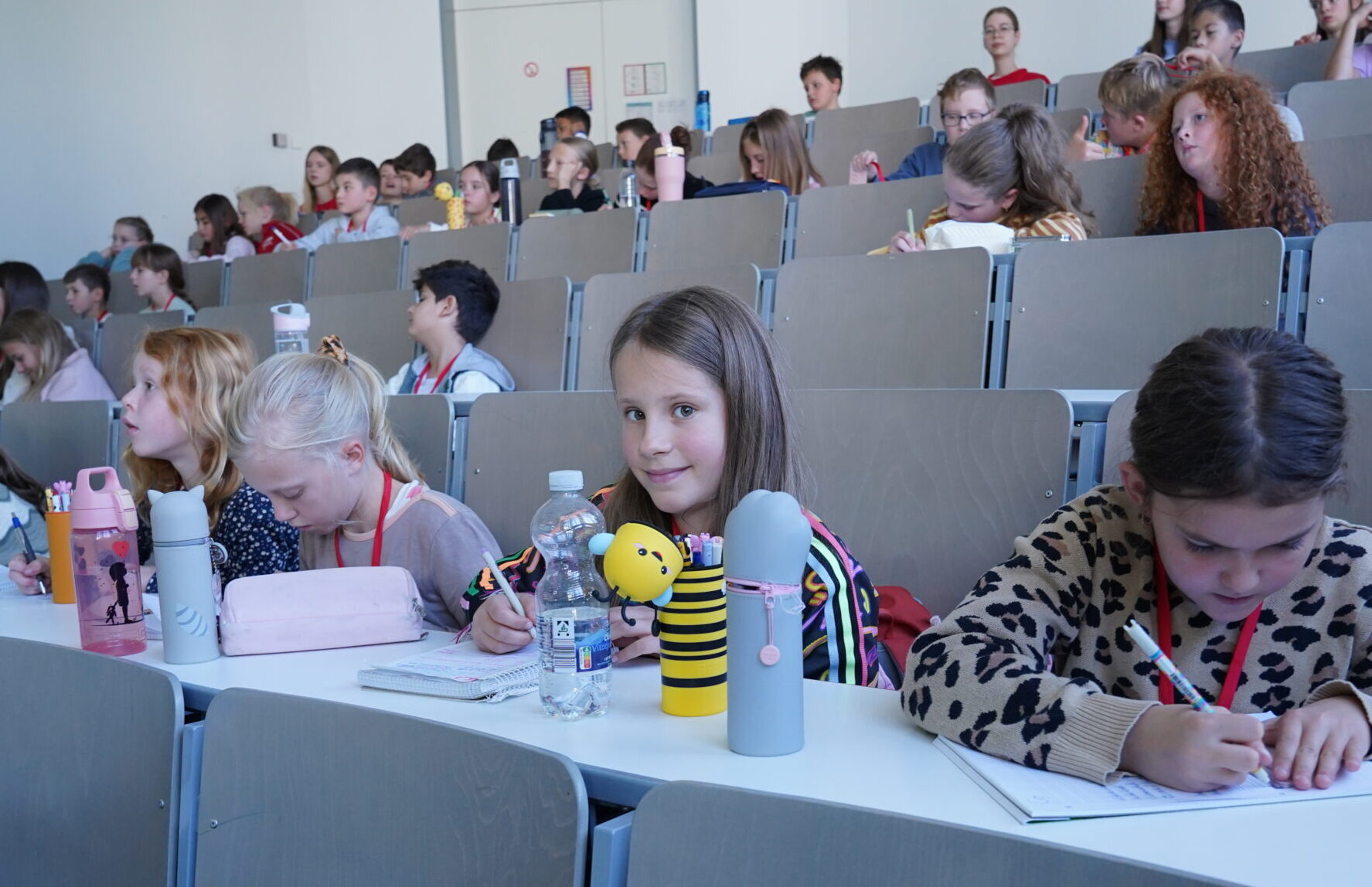 Kinder sitzen bei einer „KinderUni“-Veranstaltung an Schreibtischen in einem stufenförmig angeordneten Hörsaal der Universität; einige blicken in die Kamera, andere machen sich Notizen in ihren Heften. at Hochschule Coburg