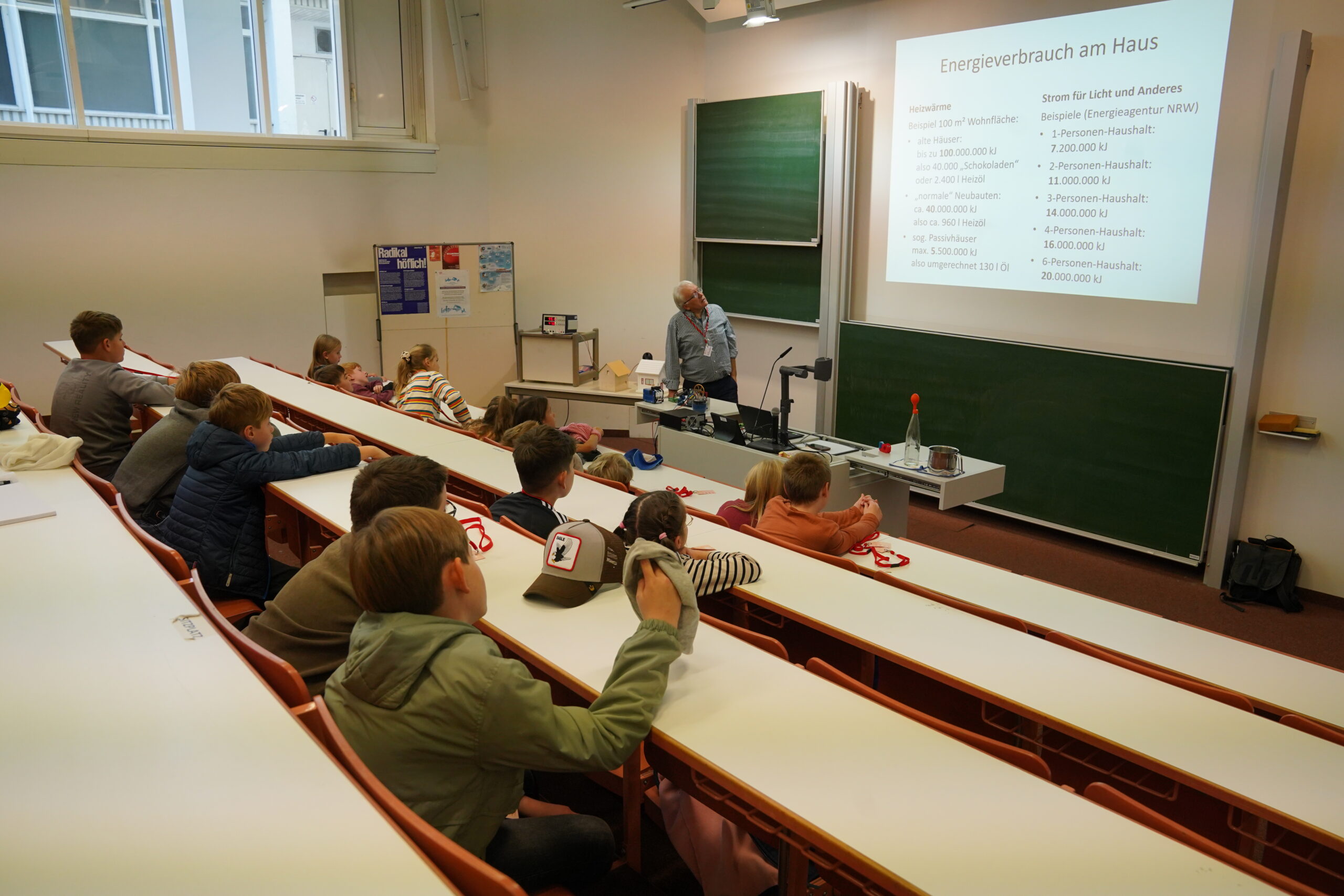 Ein Dozent steht vorne im Unterrichtsraum der KinderUni und zeigt auf eine projizierte Folie zum Thema Energieverbrauch im Haushalt, die für die Stammgäste bestimmt ist. at Hochschule Coburg