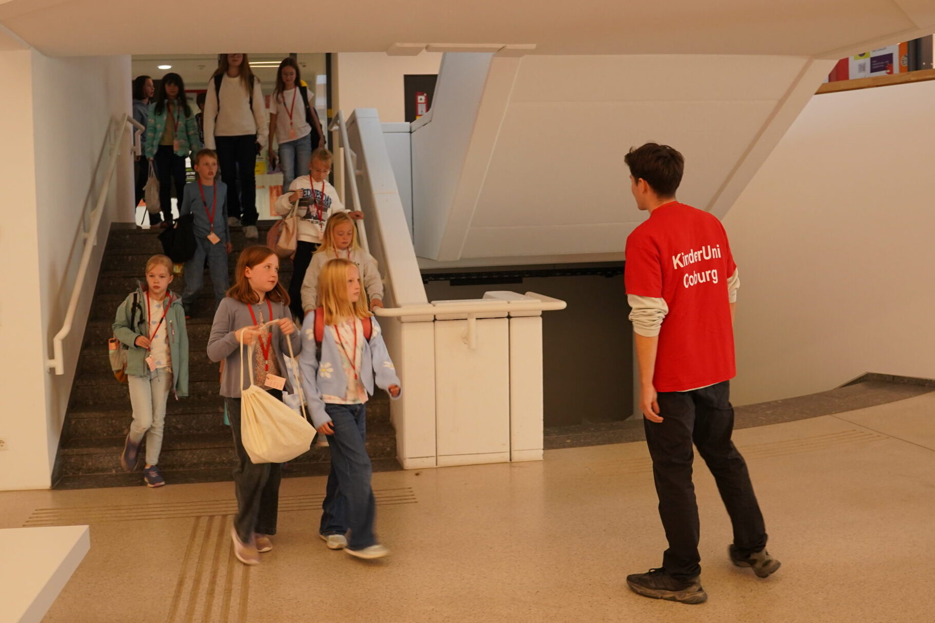 Ein Mitarbeiter in einem roten „KinderUni Coburg“-T-Shirt begrüßt eine Gruppe junger Stammgäste, die die Treppe hinunterkommt, um zur KinderUni zu gehen. at Hochschule Coburg