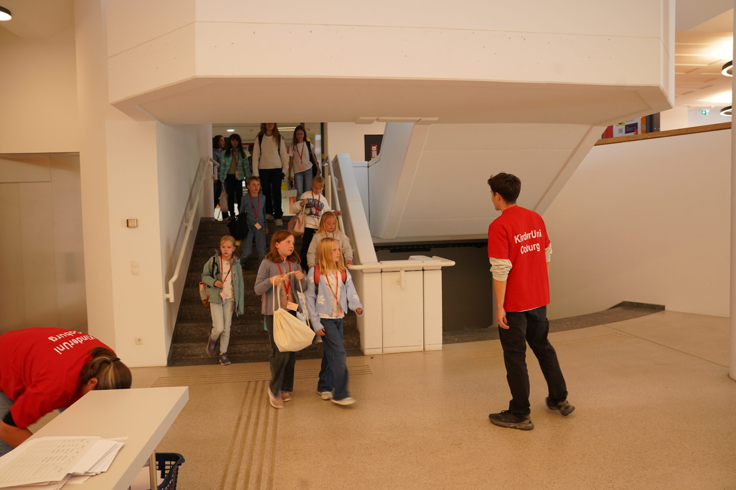 Ein Mitarbeiter in einem roten „KinderUni Coburg“-T-Shirt begrüßt eine Gruppe junger Stammgäste, die die Treppe hinunterkommt, um zur KinderUni zu gehen. at Hochschule Coburg