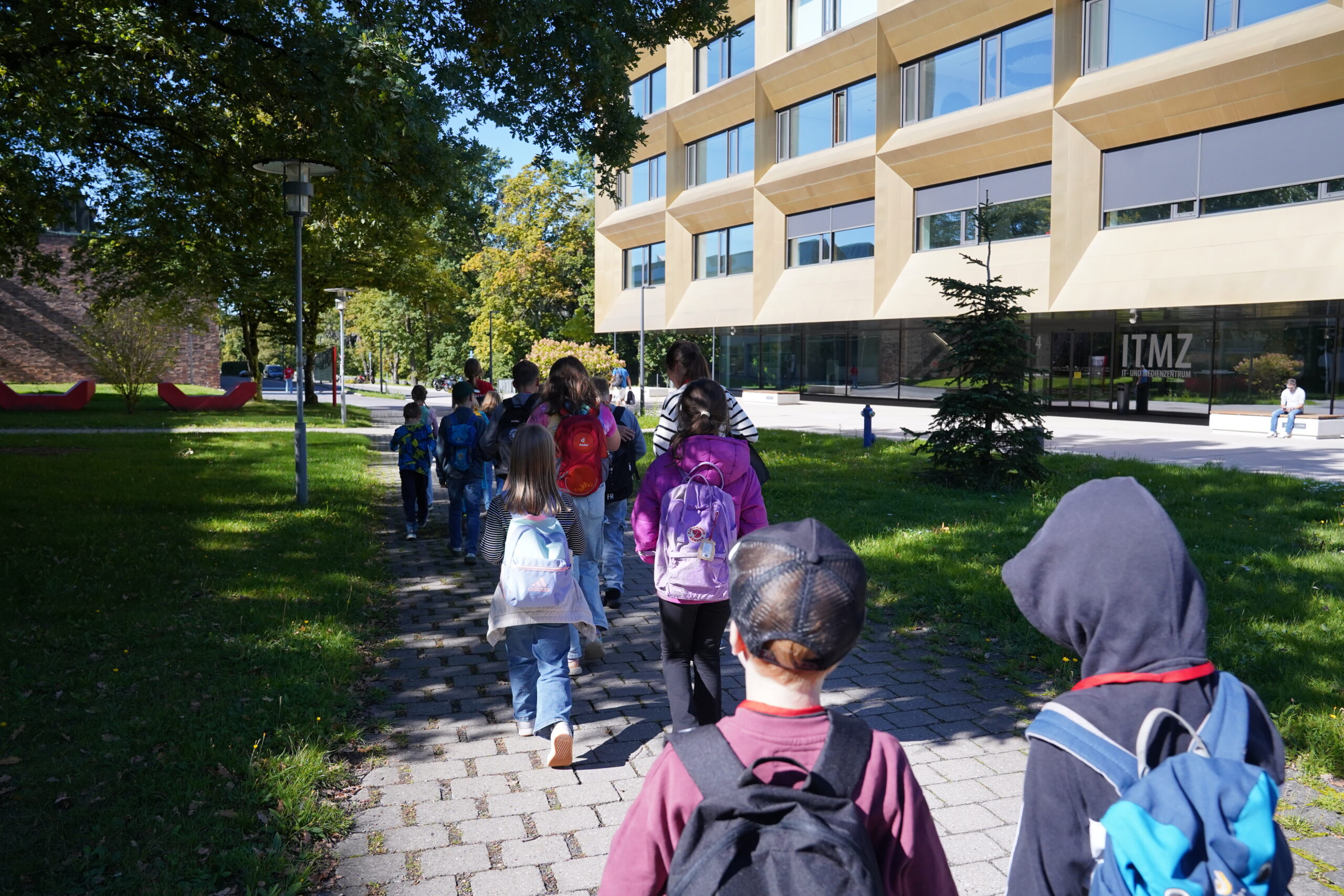 Eine Gruppe von KinderUni-Kindern mit Rucksäcken geht auf einem gepflasterten Weg auf ein modernes Universitätsgebäude mit der Aufschrift „ITMZ“ zu, genau wie die Stammgäste. at Hochschule Coburg