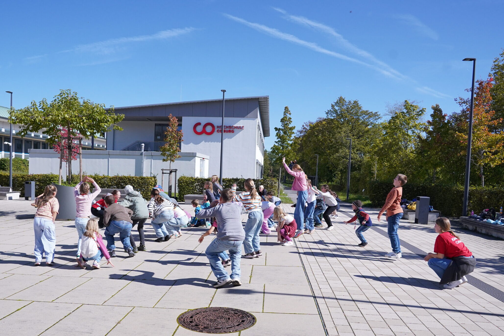 Eine Gruppe von Stammgästen, darunter Kinder und Erwachsene, macht während einer KinderUni-Veranstaltung auf einem gepflasterten Schulhof Übungen. at Hochschule Coburg
