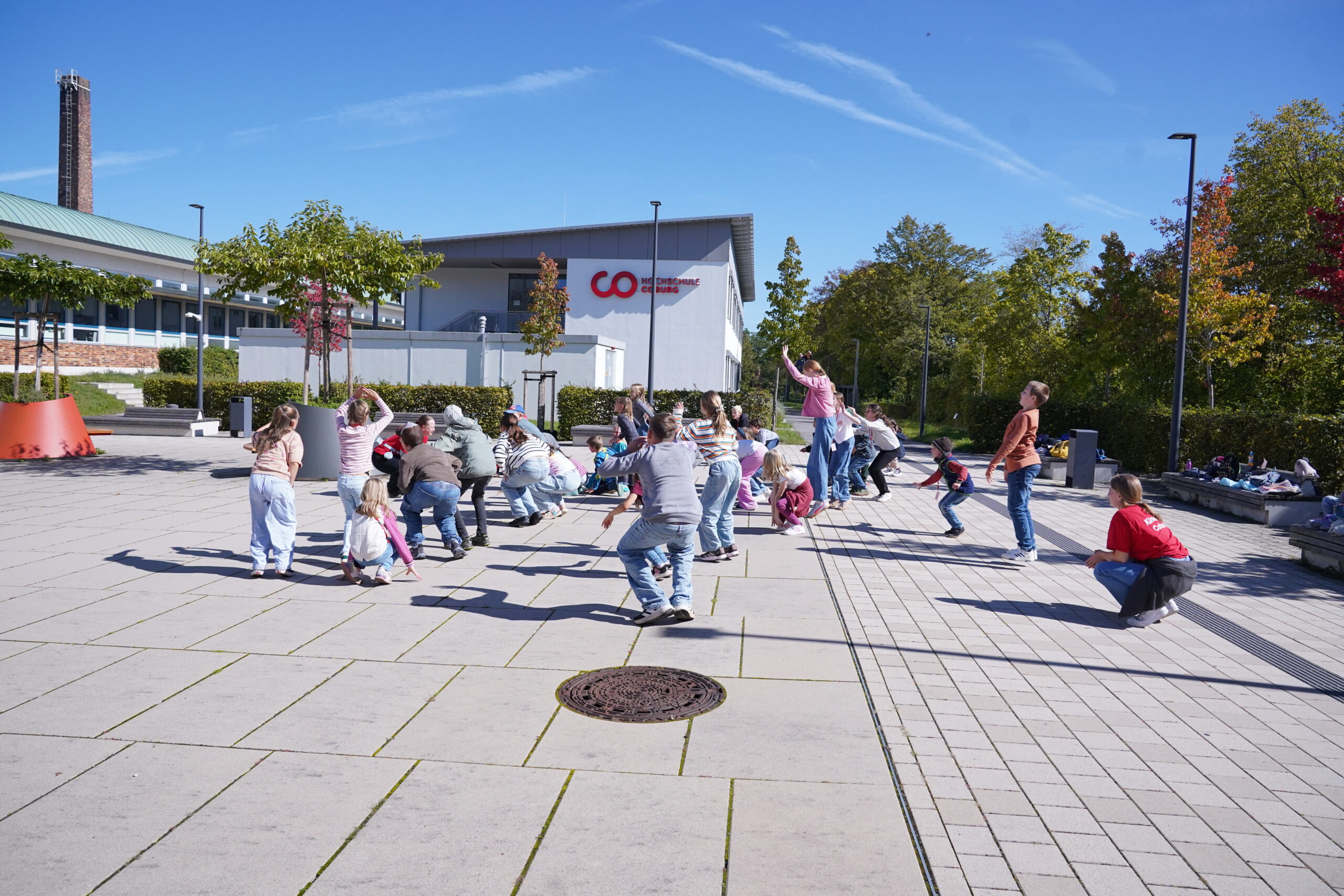 Eine Gruppe von Stammgästen, darunter Kinder und Erwachsene, macht während einer KinderUni-Veranstaltung auf einem gepflasterten Schulhof Übungen. at Hochschule Coburg
