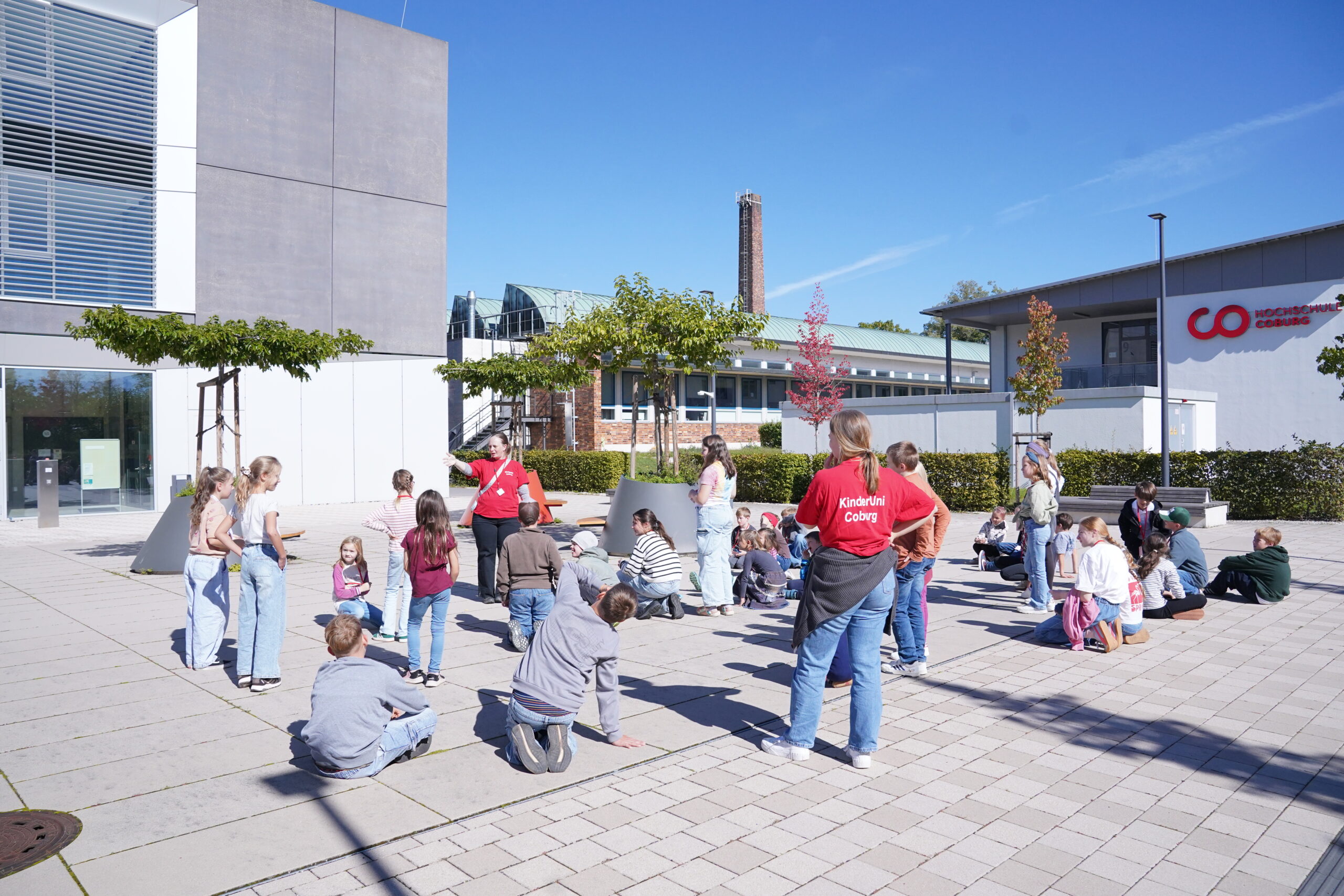 Eine große Gruppe von KinderUni-Kindern und ein Kursleiter versammeln sich auf einem gepflasterten Innenhof vor einem Universitätsgebäude, umgeben von den üblichen Stammgästen. at Hochschule Coburg
