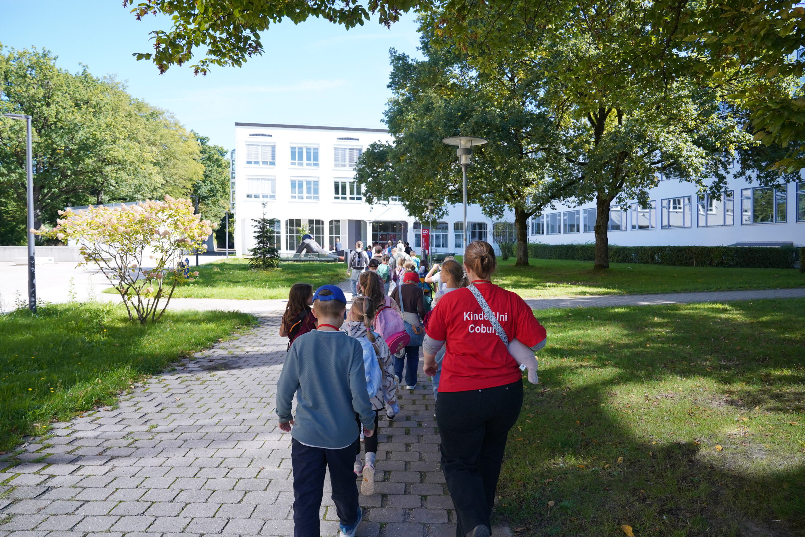 Eine Reihe von KinderUni-Teilnehmern, darunter viele Stammgäste, geht auf einem gepflasterten Weg in Richtung eines Schulgebäudes, angeführt von einem Erwachsenen in einem roten Hemd. at Hochschule Coburg