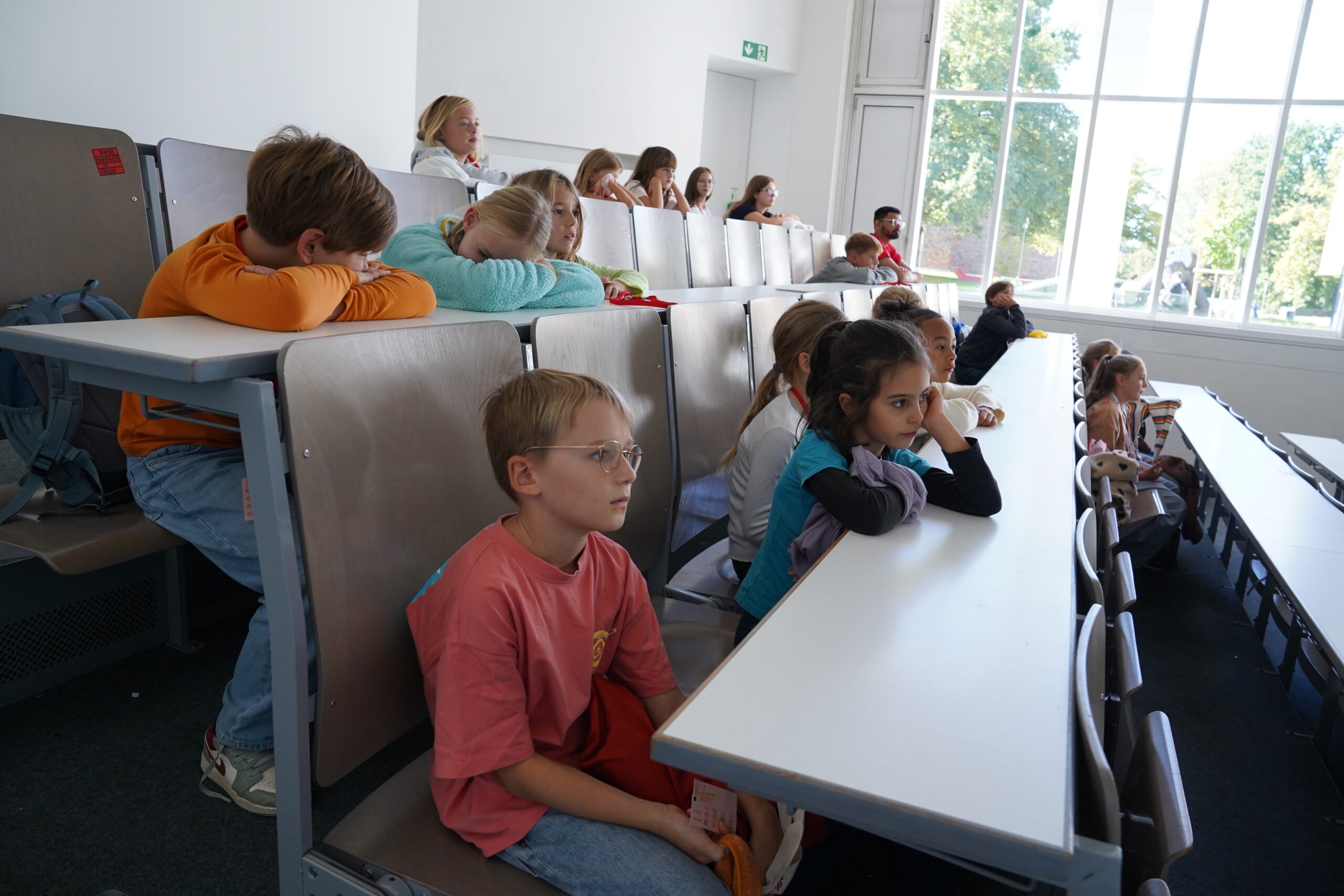 Die Studierenden sitzen an stufenförmig angeordneten Tischen in einem hell erleuchteten Hörsaal der KinderUni, und unter ihnen sind einige bekannte Gesichter, die wie Stammgäste dabei sind. at Hochschule Coburg