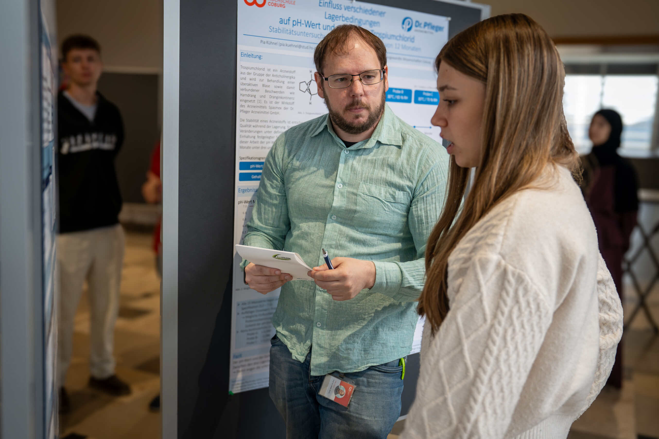 Ein Mann in einem hellgrünen Hemd bespricht auf einem wissenschaftlichen Symposium mit einer jungen Frau ein Forschungsposter von Bioanalytik. at Hochschule Coburg