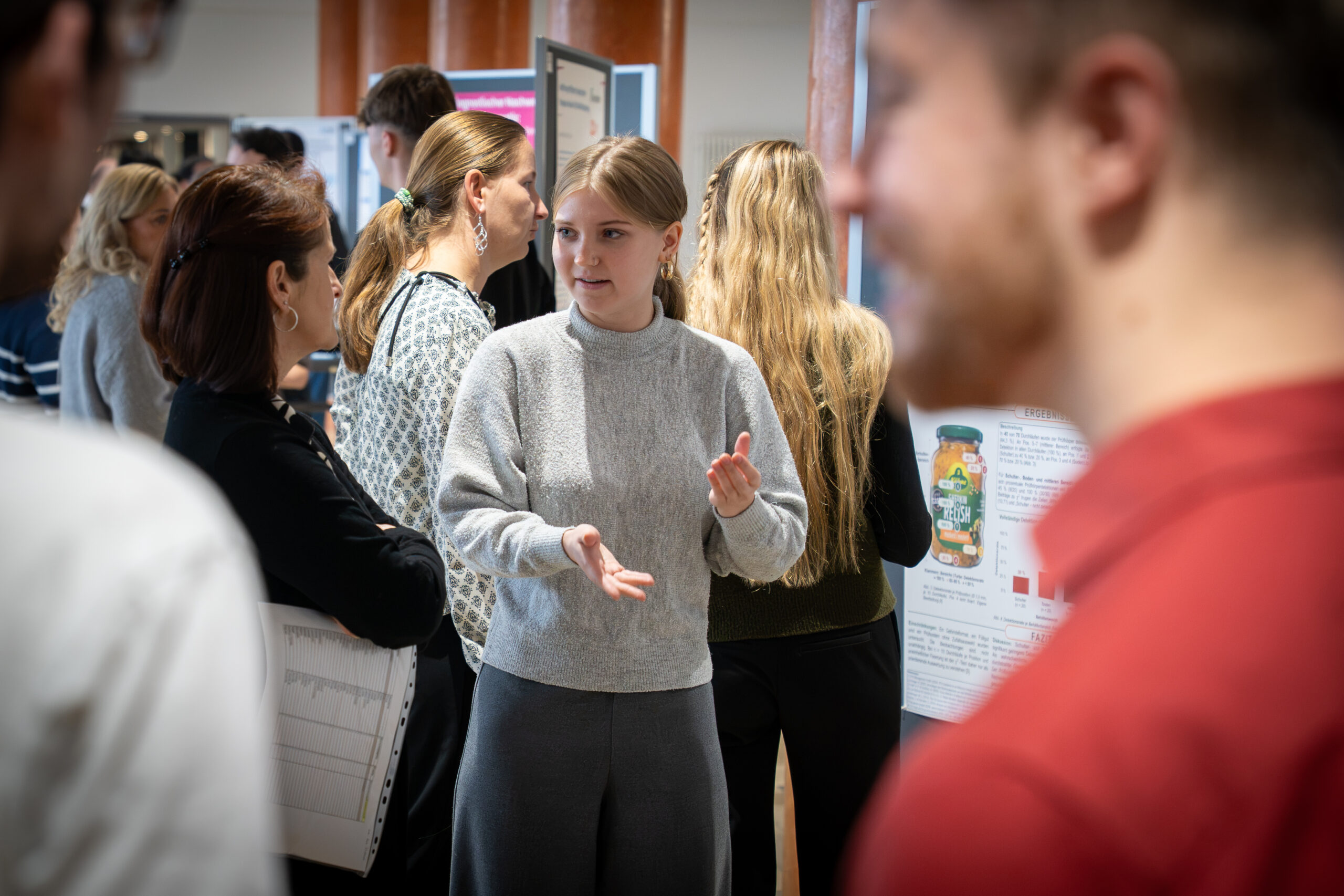 Eine junge Frau in einem grauen Pullover gestikuliert, während sie einer Gruppe, die sich vor den Forschungsplakaten auf dem Bioanalytik-Symposium versammelt hat, von ihrem Praxissemester erzählt. at Hochschule Coburg
