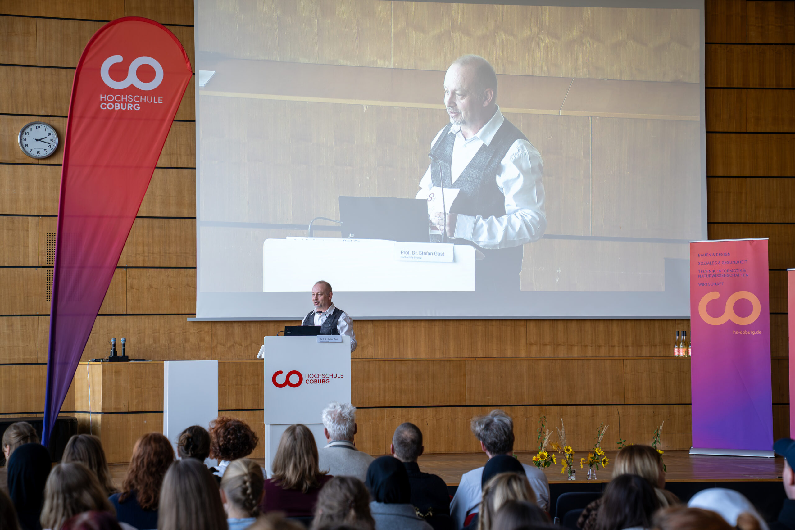 Ein Redner steht am Rednerpult des Bioanalytik-Symposiums; sein Bild wird auf eine Leinwand hinter ihm projiziert, auf der das Logo der Hochschule Coburg auf Bannern zu sehen ist. at Hochschule Coburg