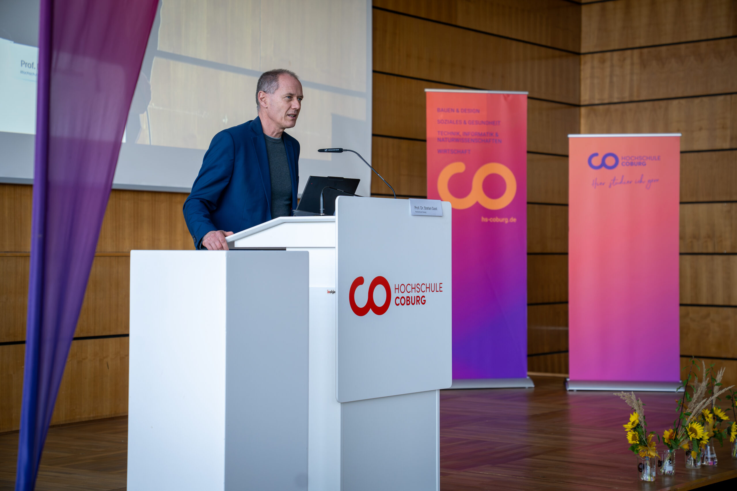Ein Mann hält auf einer Podiumsveranstaltung der Hochschule Coburg eine Rede über Bioanalytik und berufliche Perspektiven, im Hintergrund sind Banner der Hochschule zu sehen. at Hochschule Coburg
