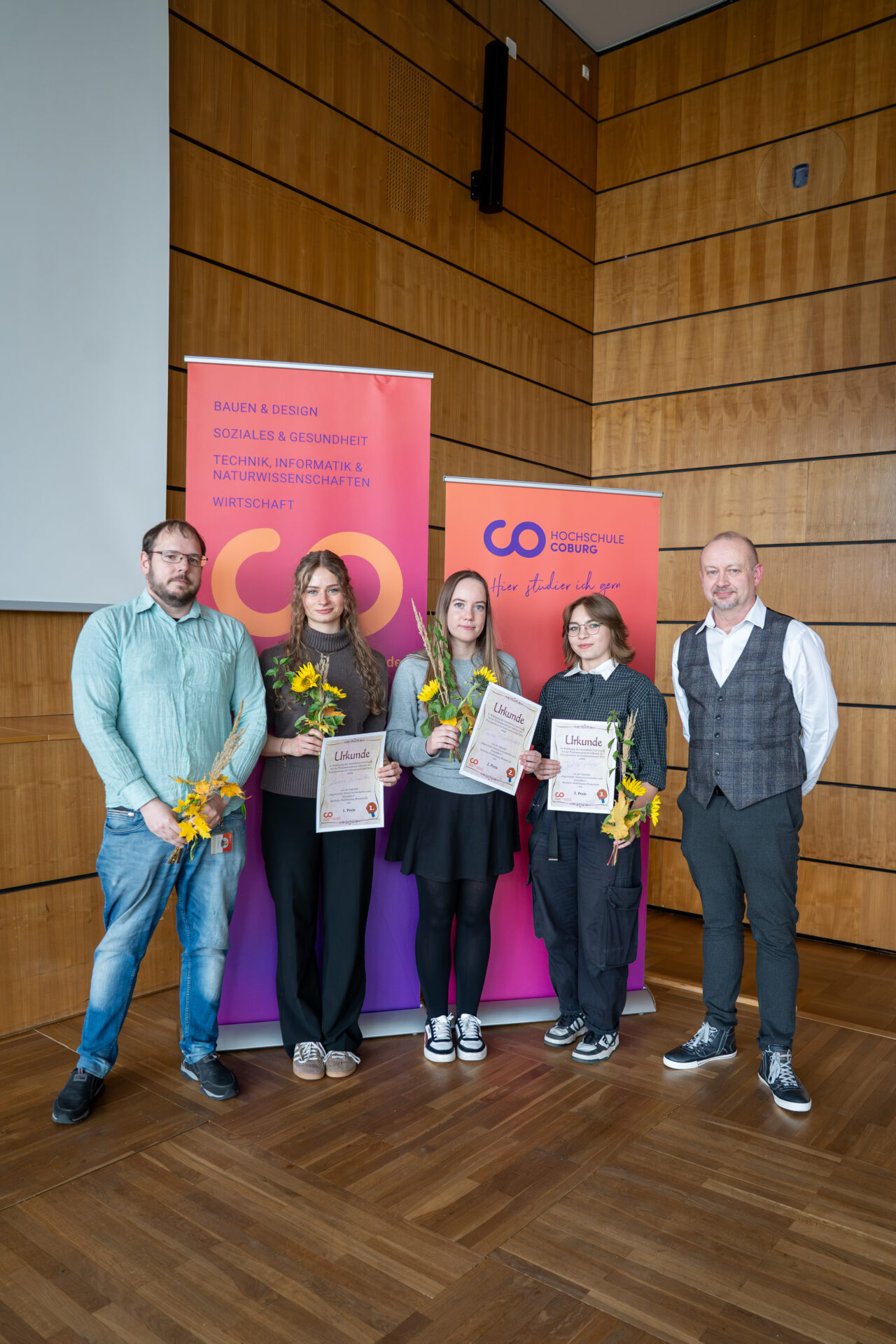 Fünf Personen stehen vor zwei Universitätsbannern; die drei Personen in der Mitte halten Urkunden und Sonnenblumen in den Händen, um ihr Praxissemester im Bereich Bioanalytik zu feiern. at Hochschule Coburg