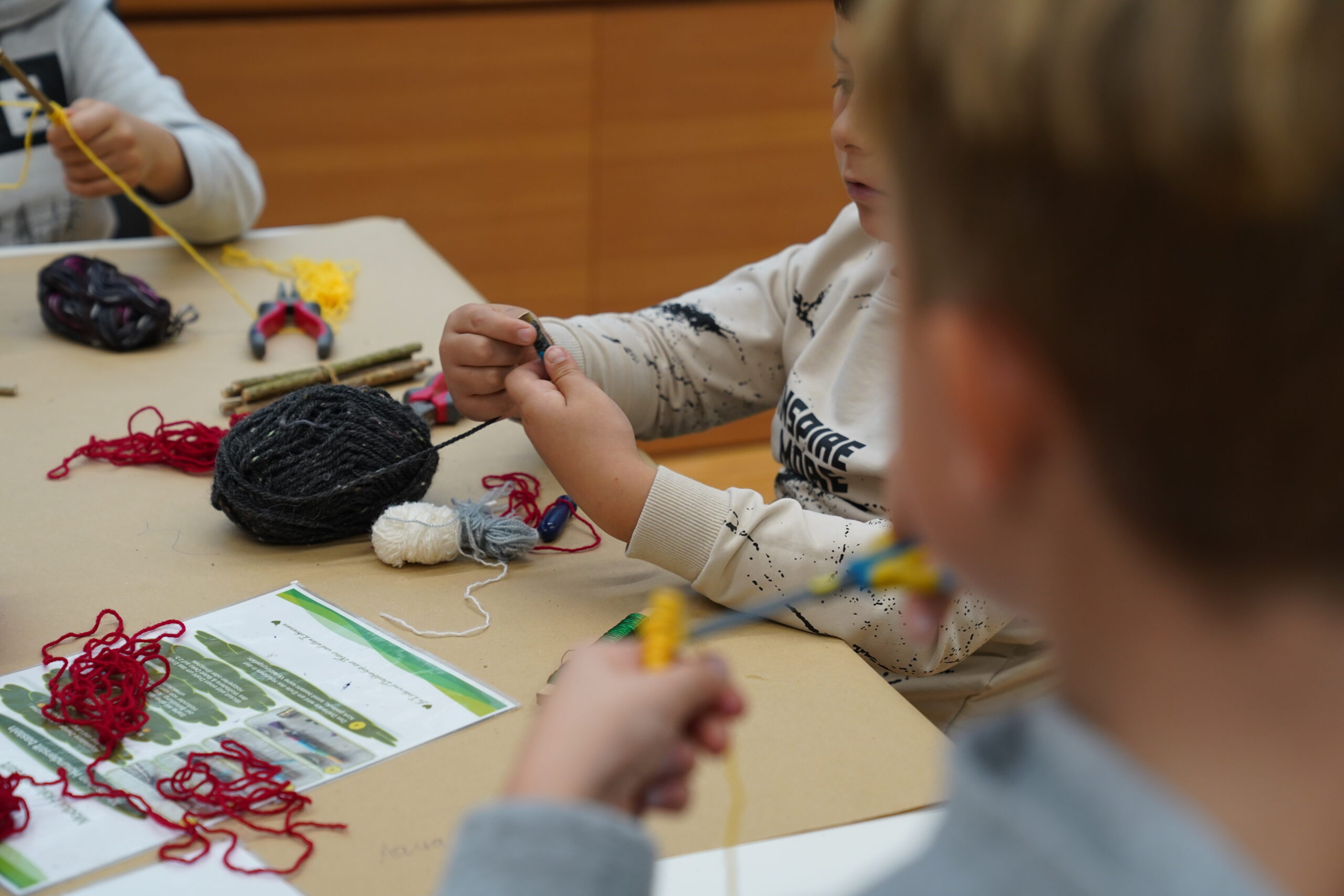 Kinder, die an einem Tisch an einem automatisierten Bastelprojekt mit Garn und kleinen Holzstäbchen arbeiten. at Hochschule Coburg