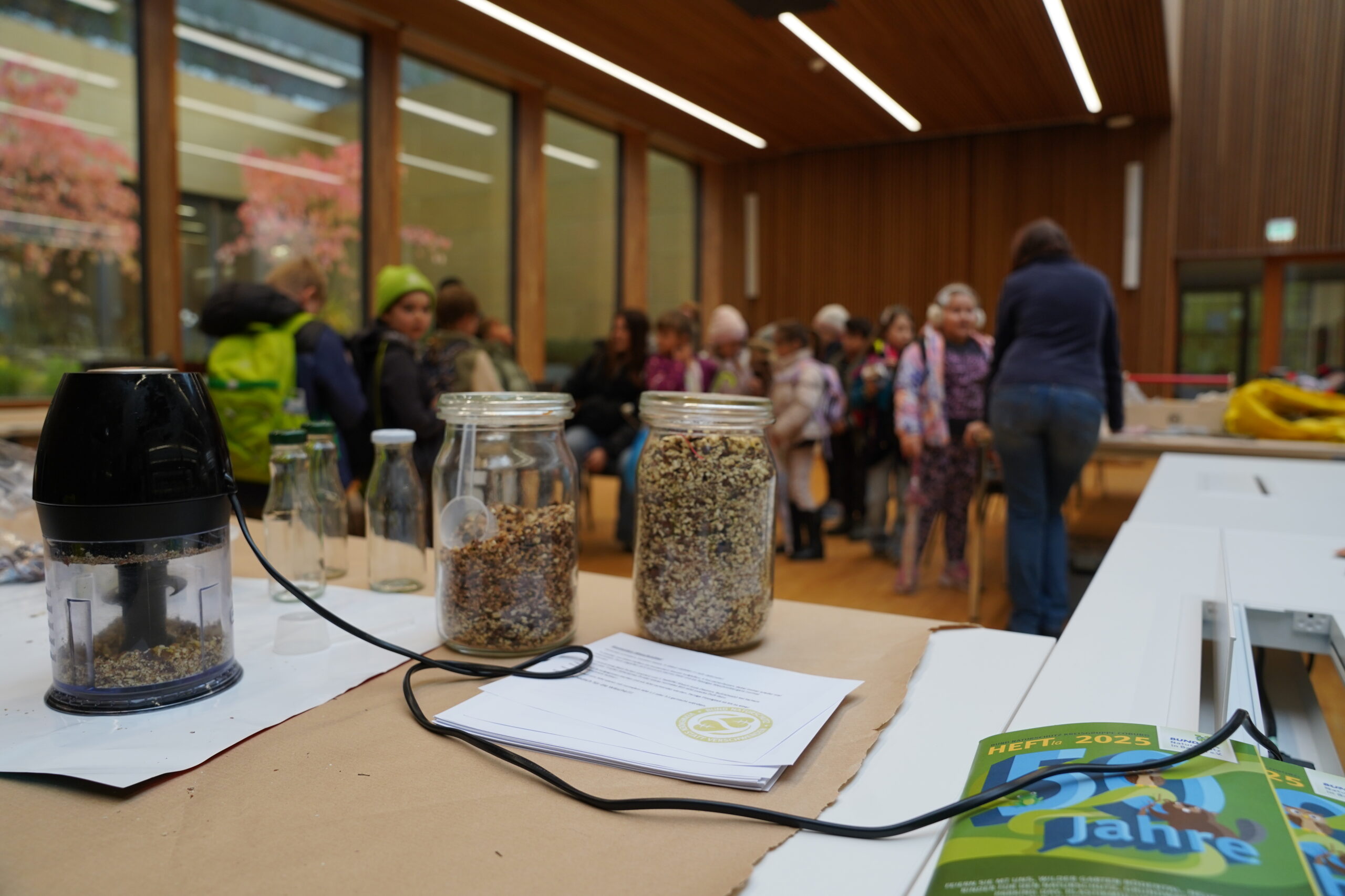 Auf einem Tisch stehen eine Küchenmaschine und zwei Gläser mit Samen, die einen automatisierten Designprozess unterstützen, während im Hintergrund eine Gruppe von Menschen zu sehen ist. at Hochschule Coburg