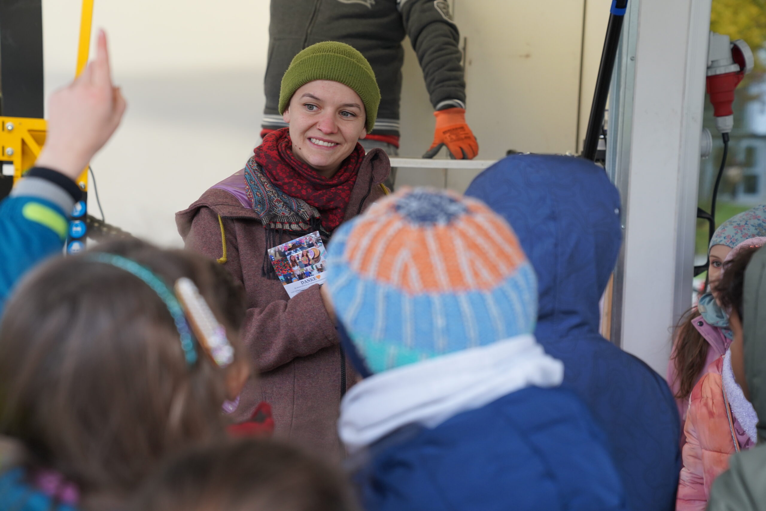 Eine lächelnde Frau mit einer grünen Mütze und einem weinroten Schal reicht einer Gruppe von Kindern eine kleine, automatisch gestaltete Fotokarte. at Hochschule Coburg