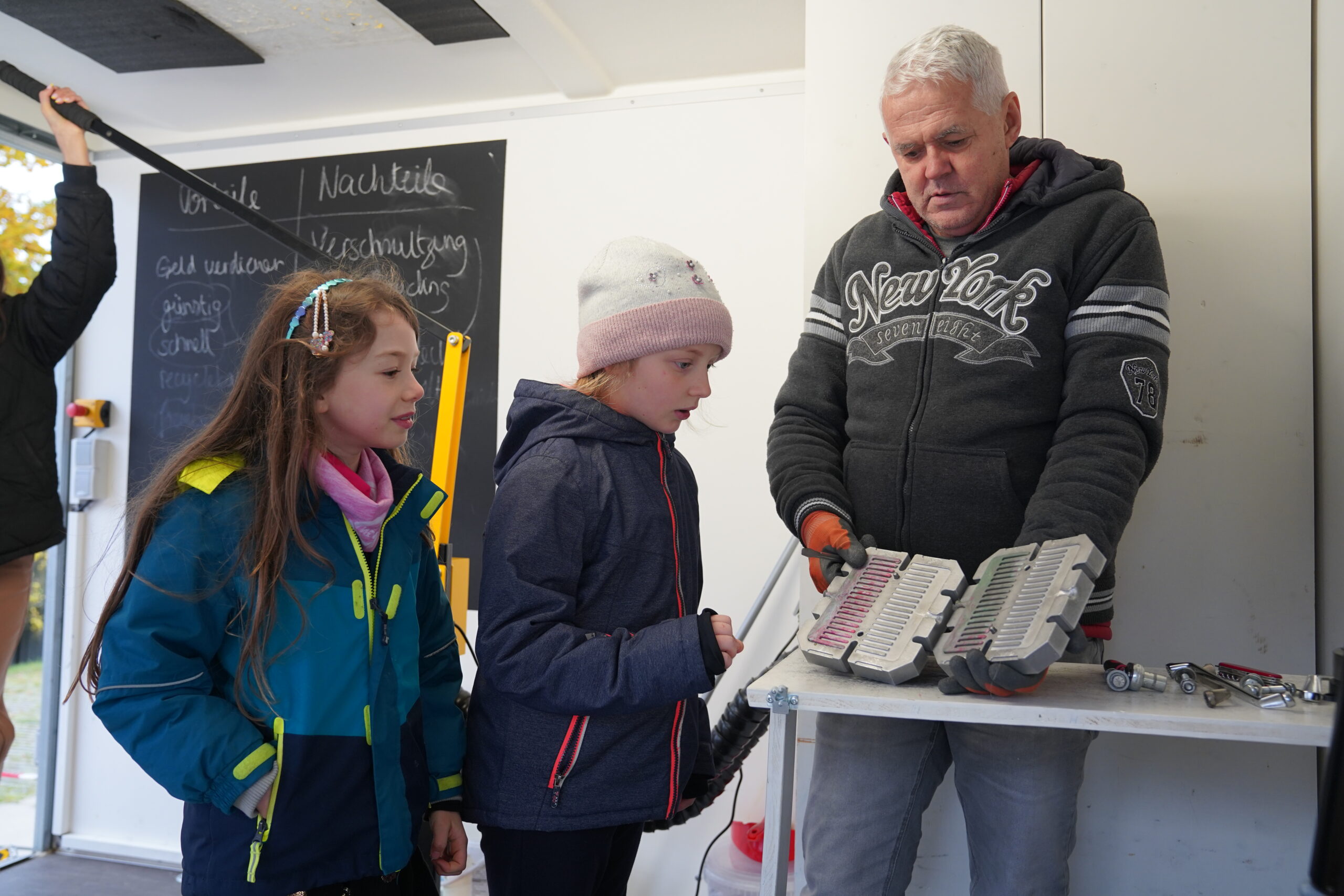 Ein älterer Mann zeigt zwei jungen Mädchen auf einer Werkbank in der Werkstatt ein Paar Metallformen und erklärt ihnen den Entstehungsprozess ihres Entwurfs. at Hochschule Coburg