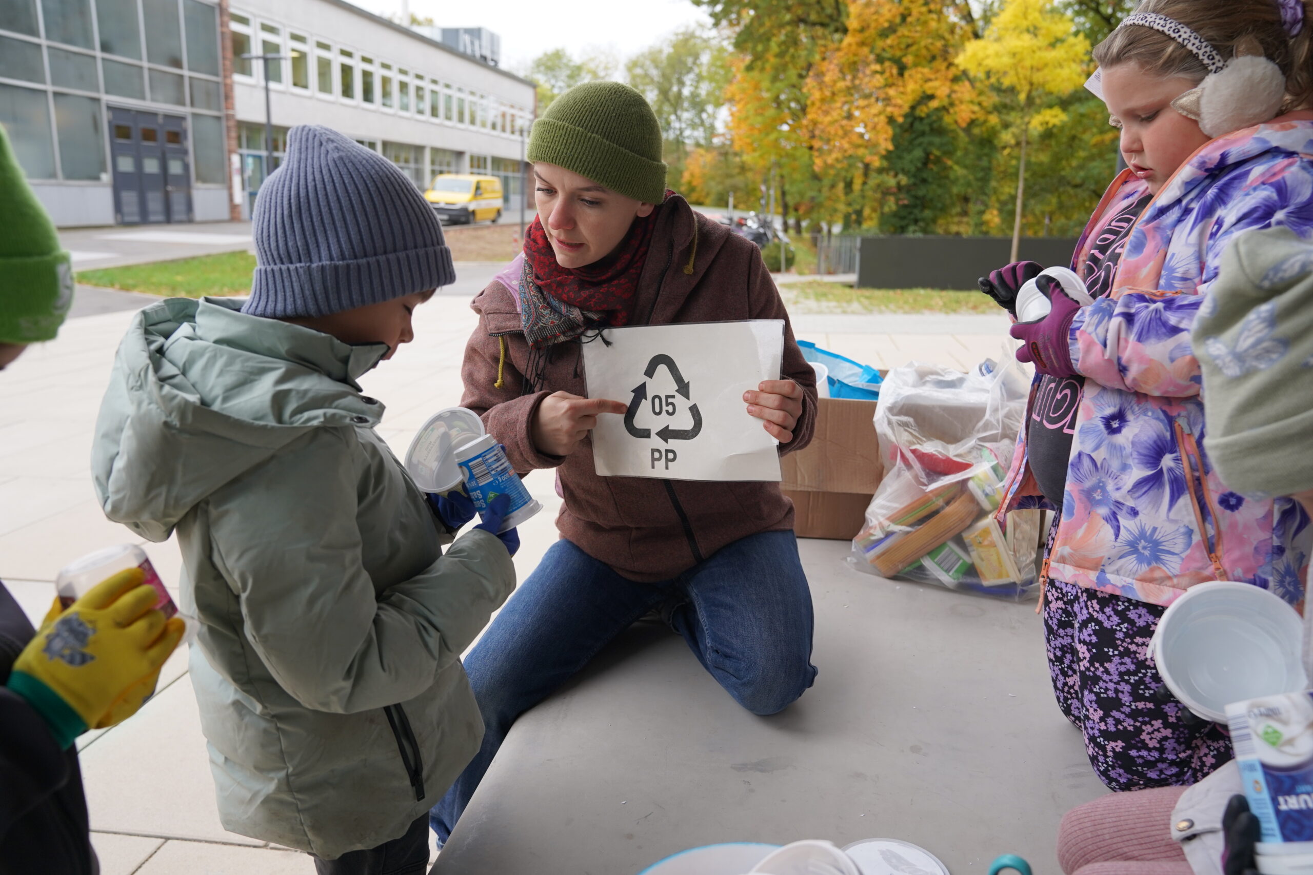 Ein Erwachsener erklärt zwei Kindern, die im Rahmen einer „automatik-entwurf“-Vorführung im Freien Plastikbehälter sortieren, das Recycling-Symbol „05 PP“. at Hochschule Coburg