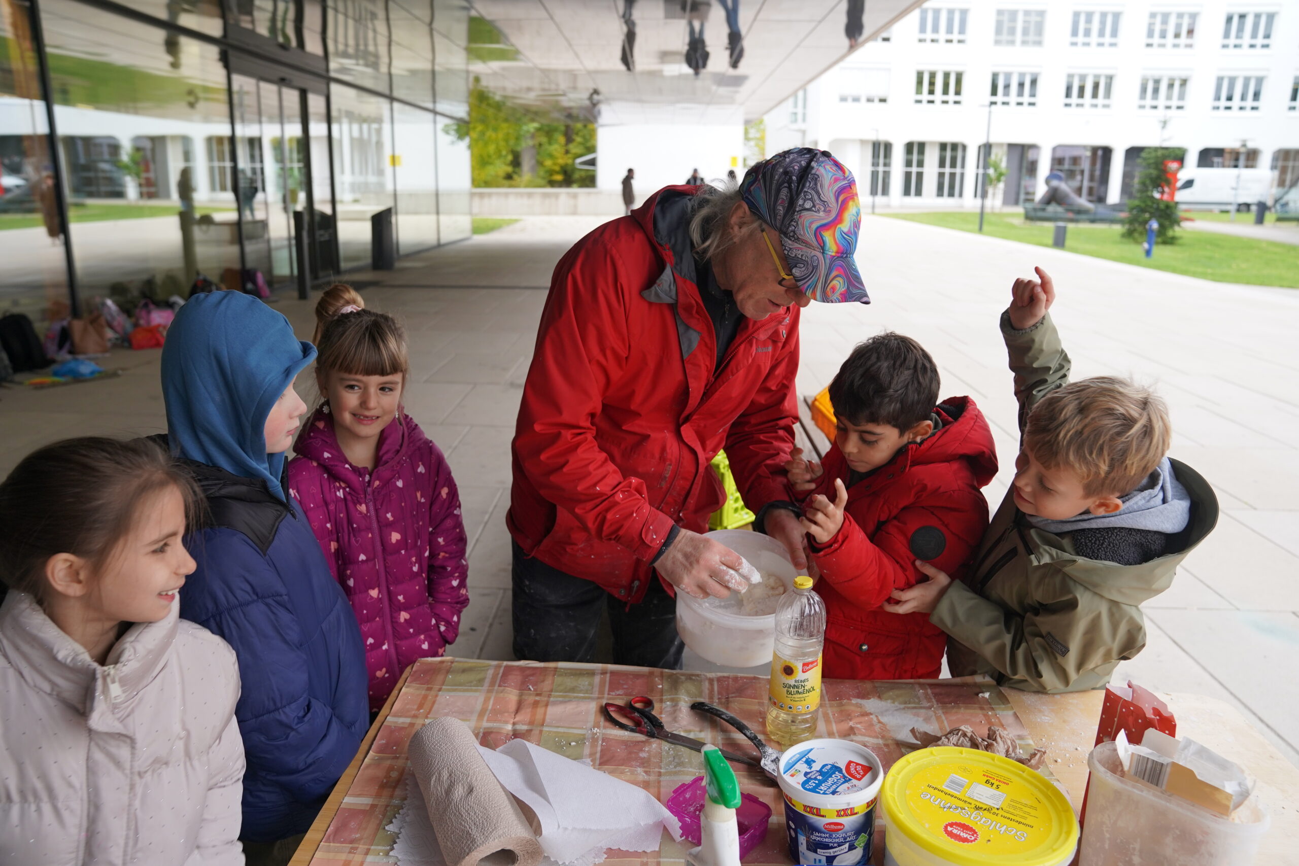 Ein Erwachsener in einer roten Jacke demonstriert einer Gruppe von Kindern an einem Tisch im Freien, wie der Teig in einer Plastikschüssel automatisch geknetet wird. at Hochschule Coburg