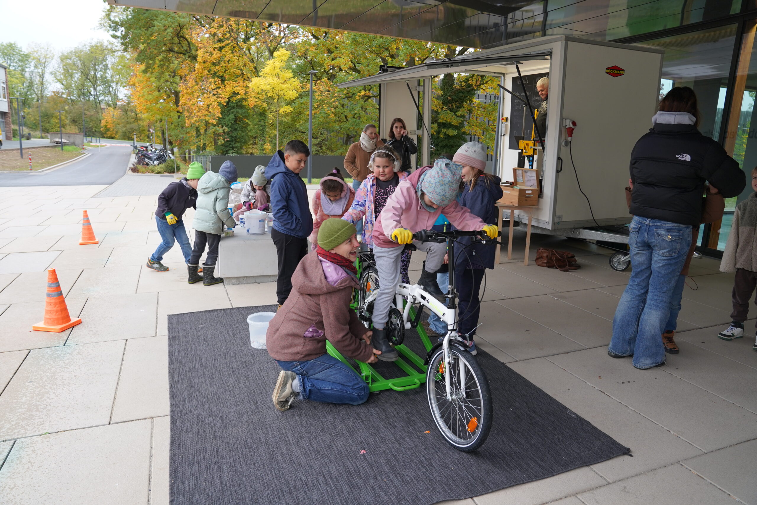 Ein Erwachsener kniet sich hin, um ein Klappfahrrad für ein Kind festzuhalten, während sich andere Kinder um einen mobilen Werkstattanhänger mit einem automatisierten „Entwurf“-Design versammeln. at Hochschule Coburg