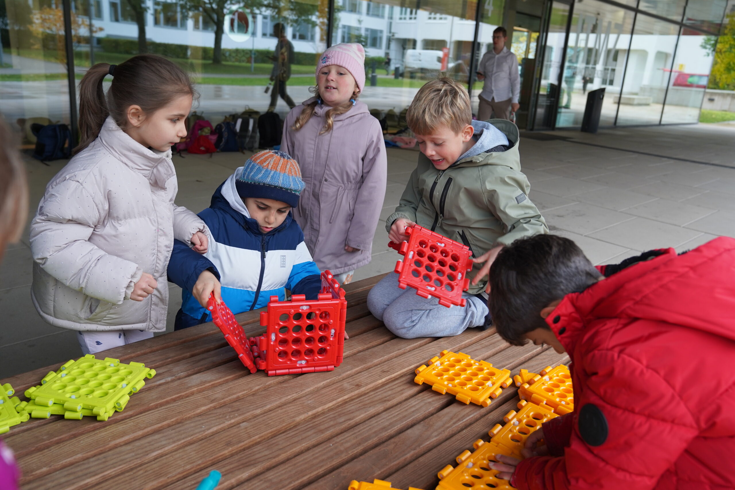 Fünf Kinder sitzen an einem Holztisch im Freien und nutzen bunte Plastikbauklötze für einen automatischen Entwurf. at Hochschule Coburg