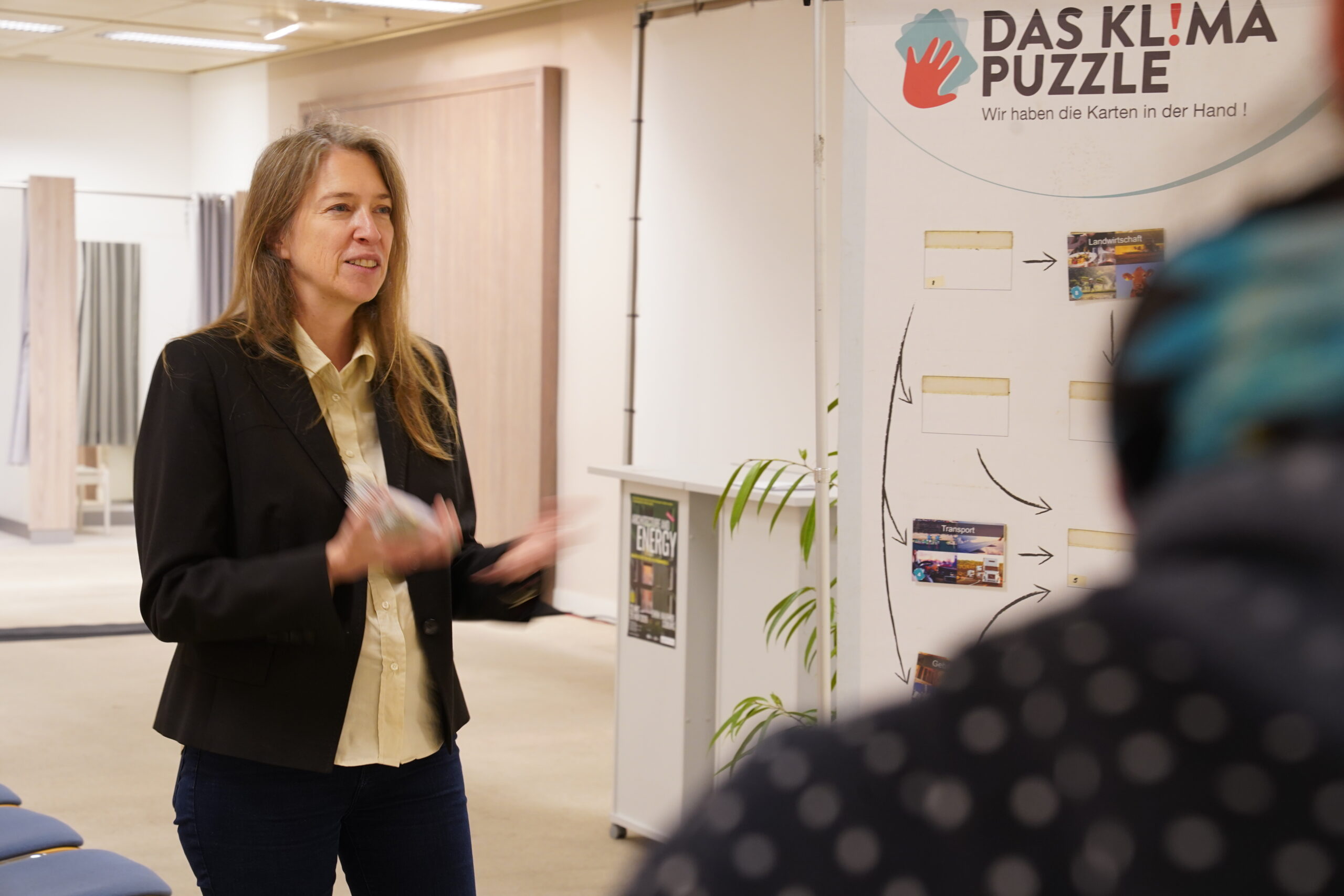 Eine Frau gestikuliert, während sie neben einer pädagogischen Ausstellung mit dem Titel „Das Klima-Puzzle“ spricht, die ein automatisches Design aufweist. at Hochschule Coburg