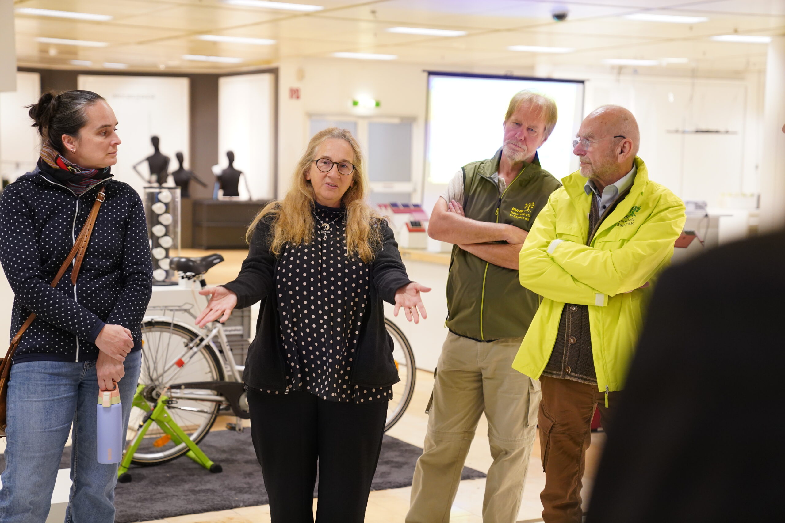 Eine Frau gestikuliert, während sie mit zwei Männern in einem hell beleuchteten Innenraum über ein automatisiertes Design spricht; im Hintergrund ist ein Fahrrad zu sehen. at Hochschule Coburg