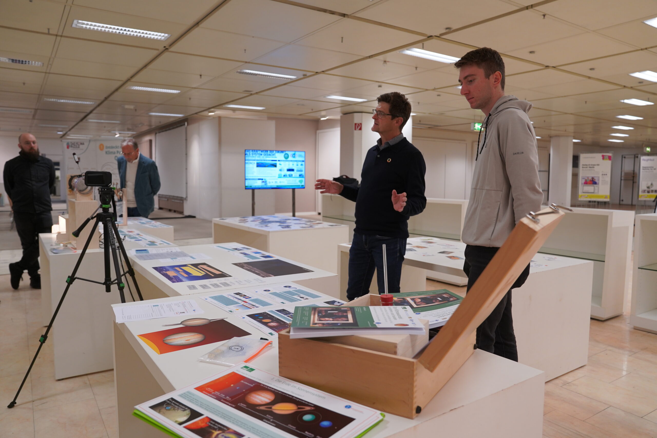 Ein Mann gestikuliert, während er einem anderen Mann in einer Ausstellungshalle einen „automatischen Entwurf“ auf Plakaten erklärt; ein dritter Mann steht daneben. at Hochschule Coburg