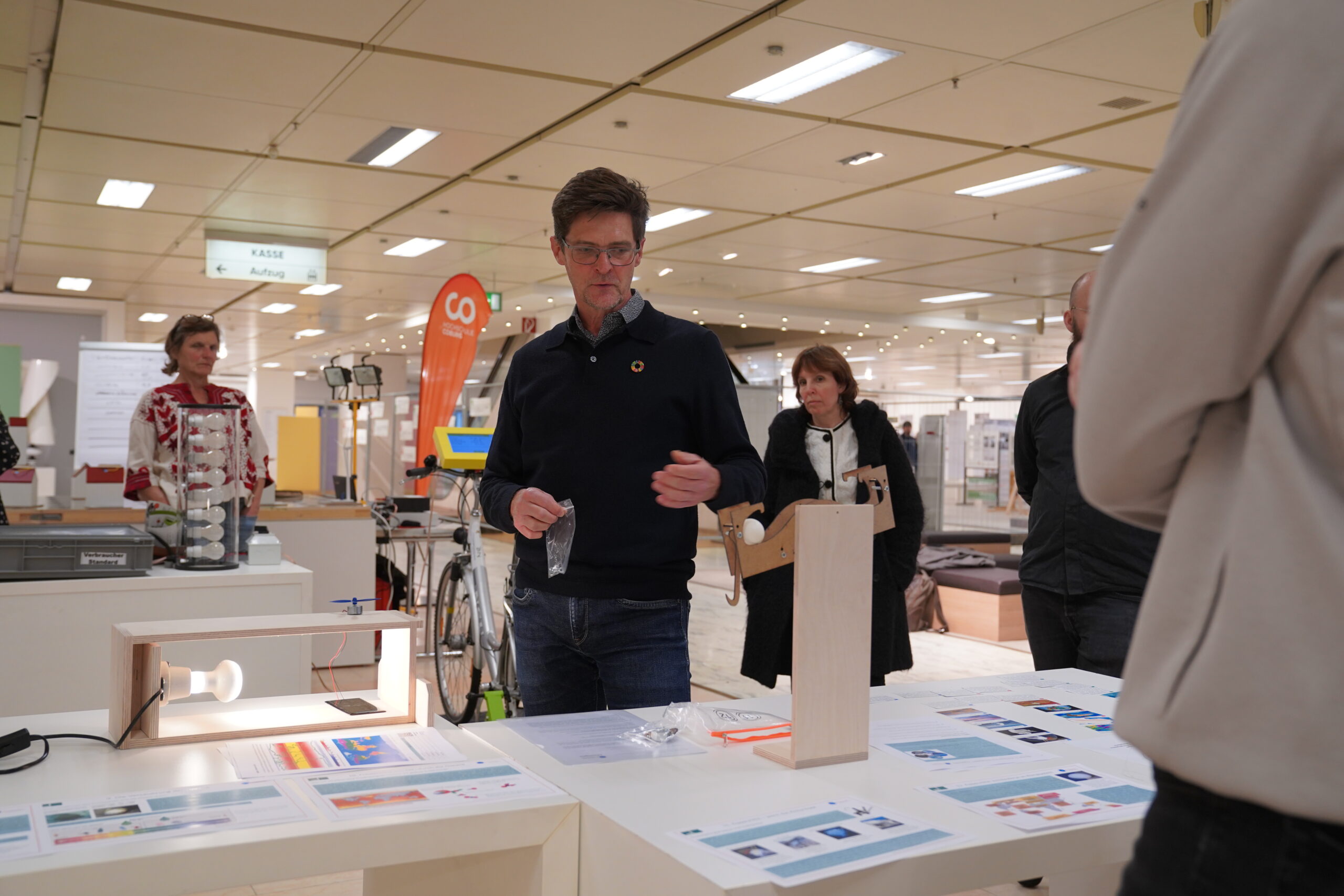 Ein Mann in einem marineblauen Pullover deutet auf eine kleine Holzleuchte – einen Prototyp für automatisiertes Design –, die auf einem Ausstellungsstand auf einer Messe steht. at Hochschule Coburg