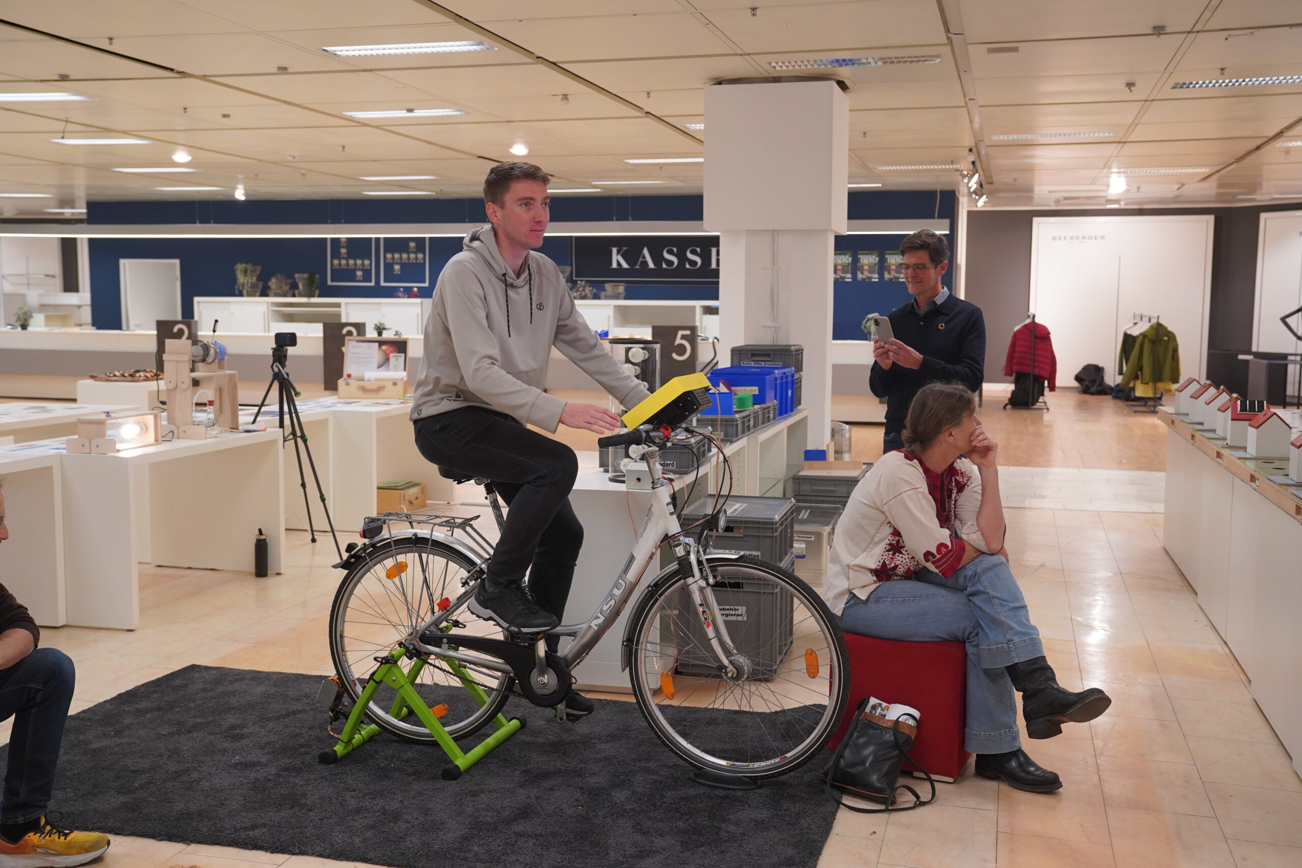 Ein Mann in einem grauen Kapuzenpullover sitzt für eine „Entwurf“-Vorführung auf einem Heimtrainer, während ein Mann ihn fotografiert und eine Frau in der Nähe auf einem roten Hocker in einer Ausstellungshalle sitzt. at Hochschule Coburg