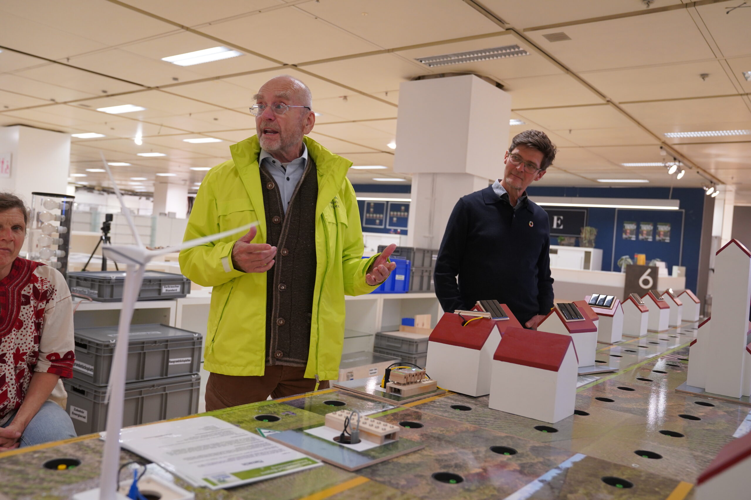 Ein Mann in einer leuchtend gelben Jacke gestikuliert, während er über einen automatischen Entwurf einer nachhaltigen Stadt mit kleinen Häusern und einer Windkraftanlage spricht. at Hochschule Coburg