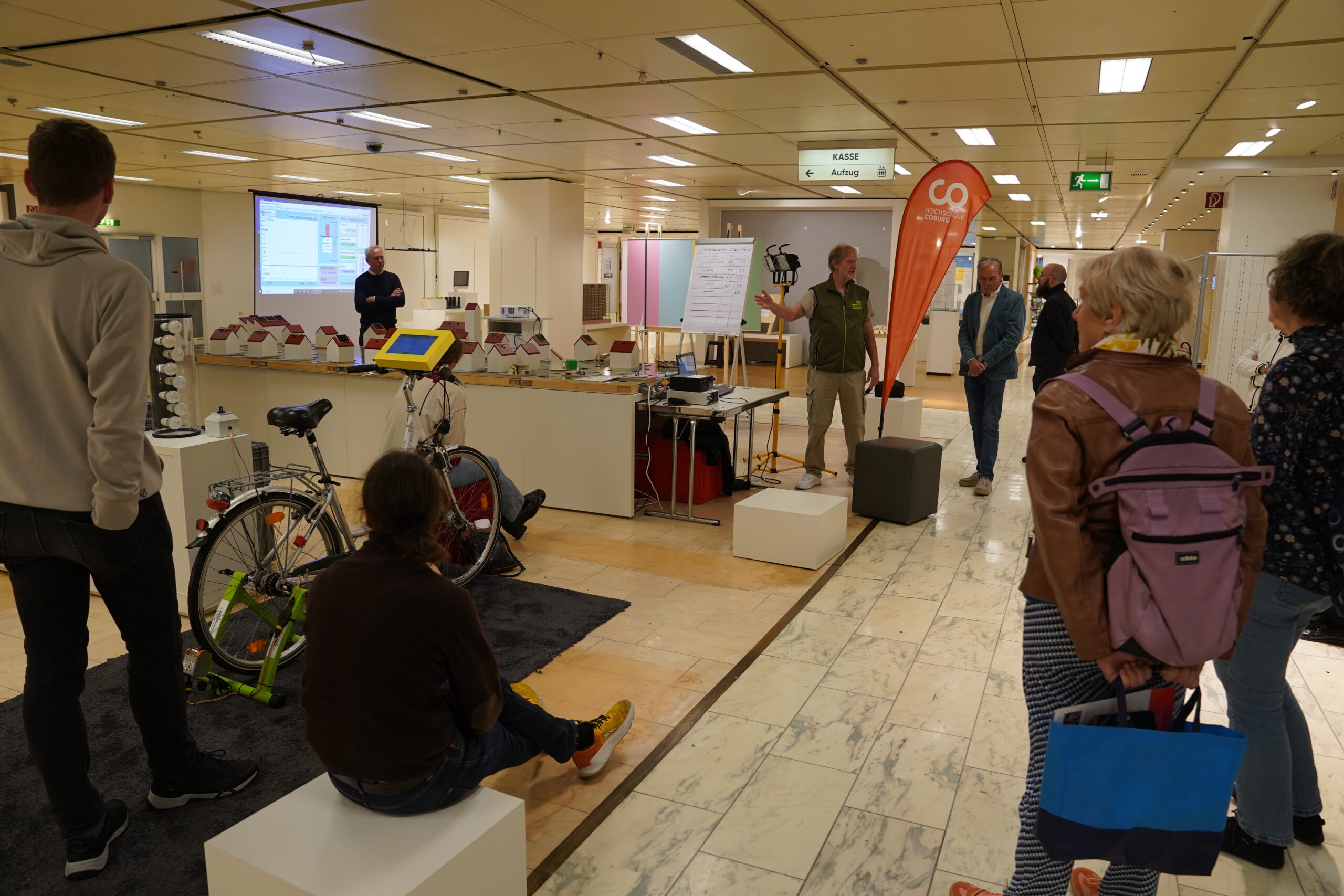 Eine Gruppe von Menschen verfolgt in einer großen Halle eine Präsentation zum Thema „automatisches Design“, bei der ein stationäres Fahrrad, eine Projektionsleinwand und ein Lautsprecher zum Einsatz kommen. at Hochschule Coburg