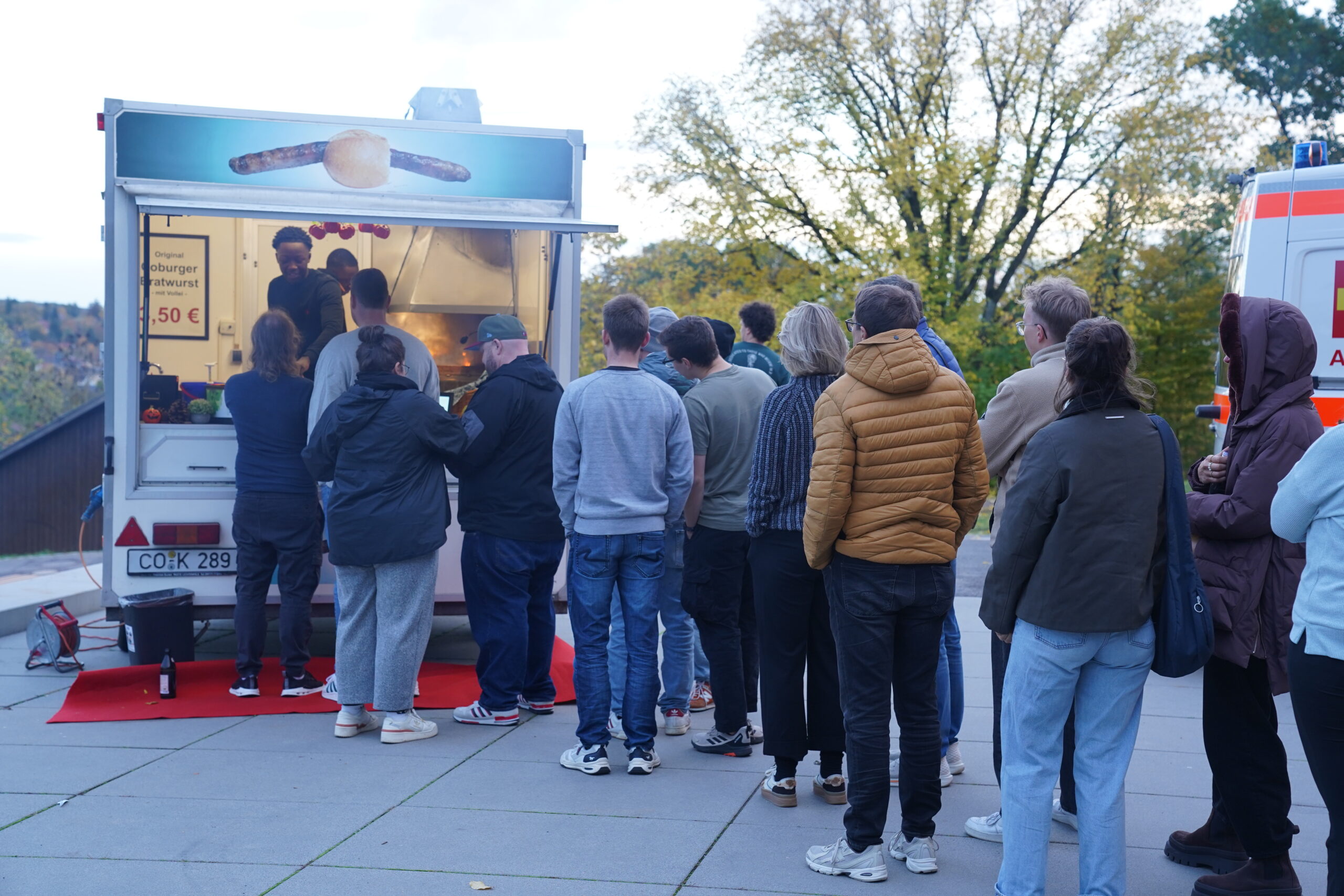 Eine Gruppe von Menschen, die in einer Schlange stehen und darauf warten, an einem Wurststand Essen zu bestellen, dargestellt als automatischer Entwurf. at Hochschule Coburg